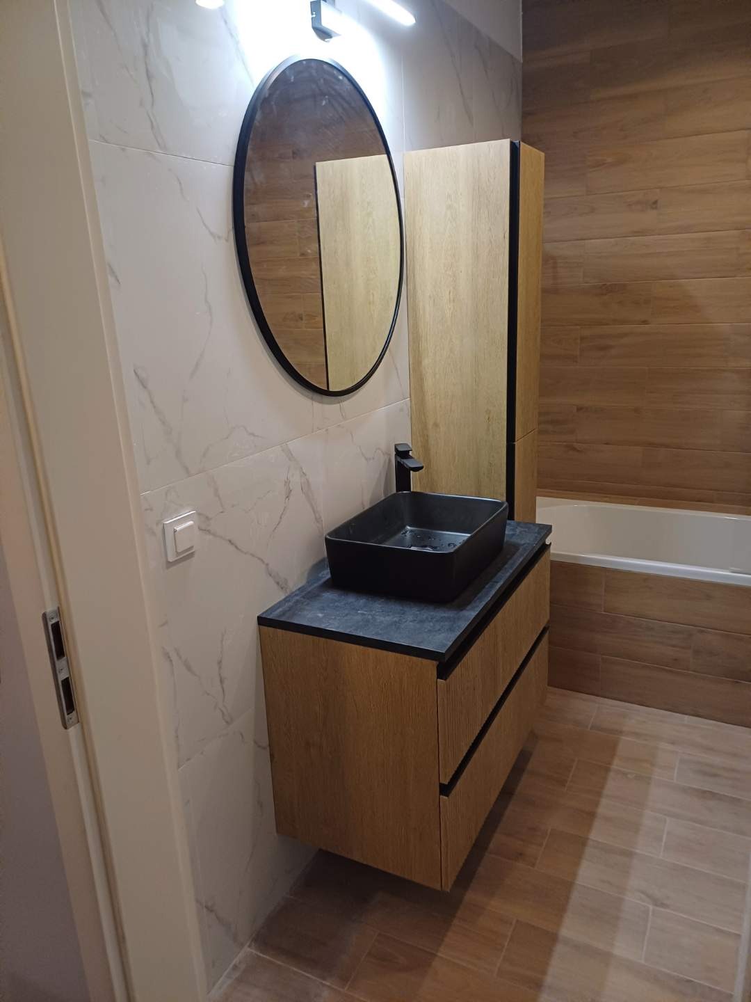 Nowoczesna łazienka z drewnopodobnymi płytkami, okrągłym lustrem i czarną umywalką nablatową na szafce z dwiema szufladami.