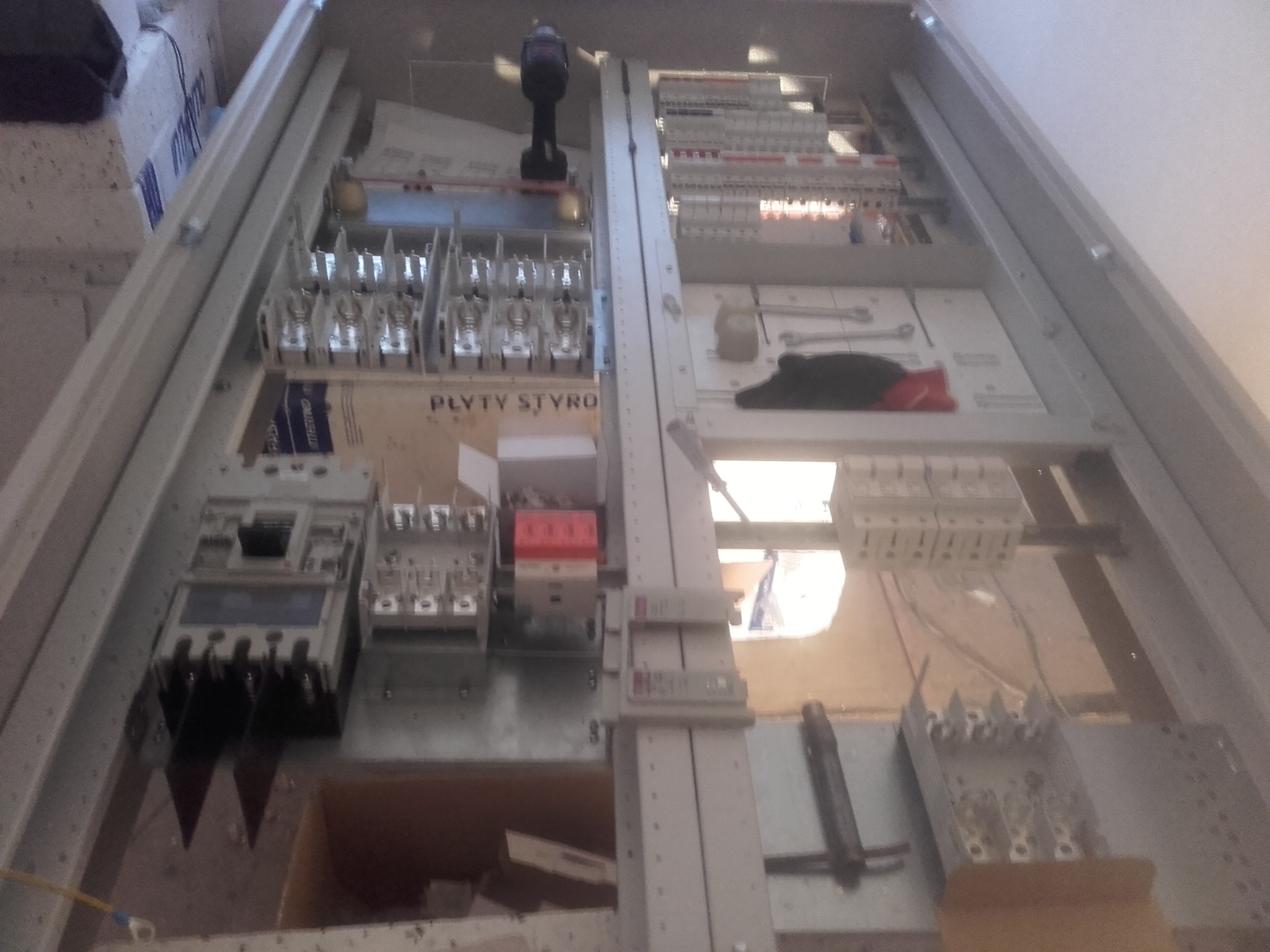 Wnętrze rozdzielnicy elektrycznej w trakcie montażu, widoczne bezpieczniki, styczniki, przewody, narzędzia i płyty styropianowe.