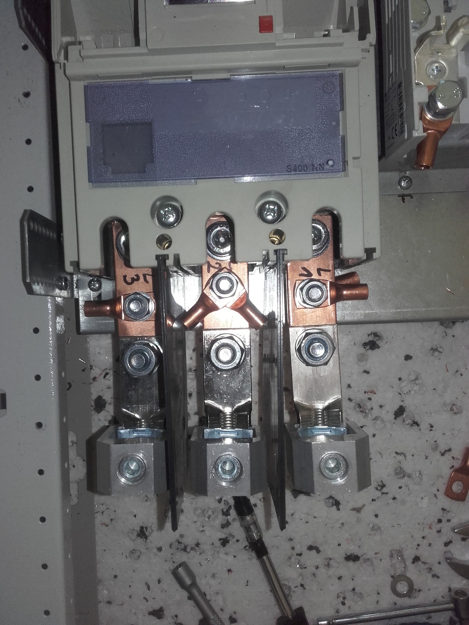 Szyna zbiorcza w rozdzielnicy elektrycznej z widocznymi przyłączami kablowymi i śrubami mocującymi, na tle zabrudzonej powierzchni roboczej.