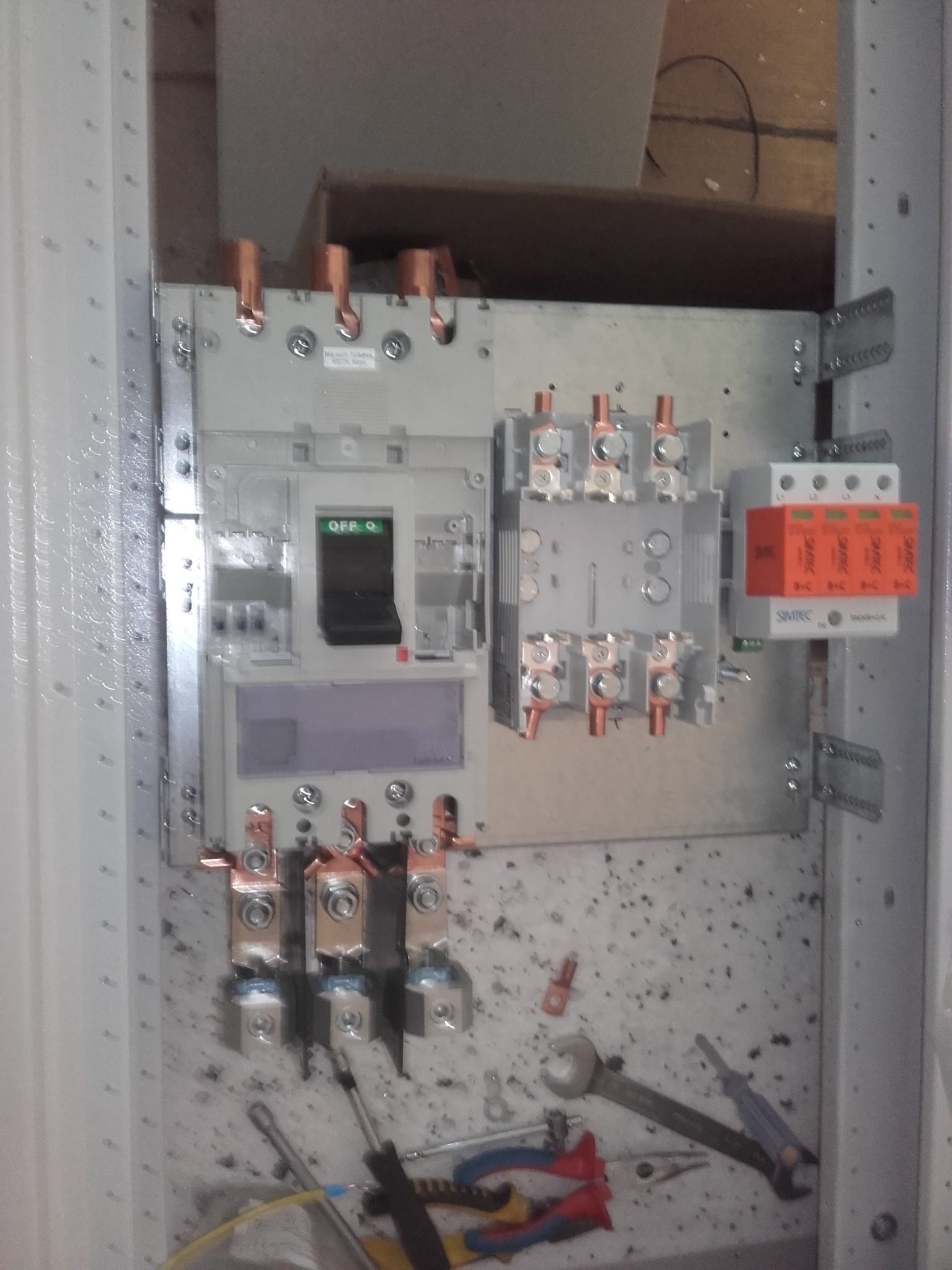 Otwarta skrzynka elektryczna z wyłącznikiem głównym w pozycji OFF, z widocznymi elementami instalacji elektrycznej, takimi jak listwy zaciskowe, ograniczniki przepięć oraz narzędzia elektryczne...