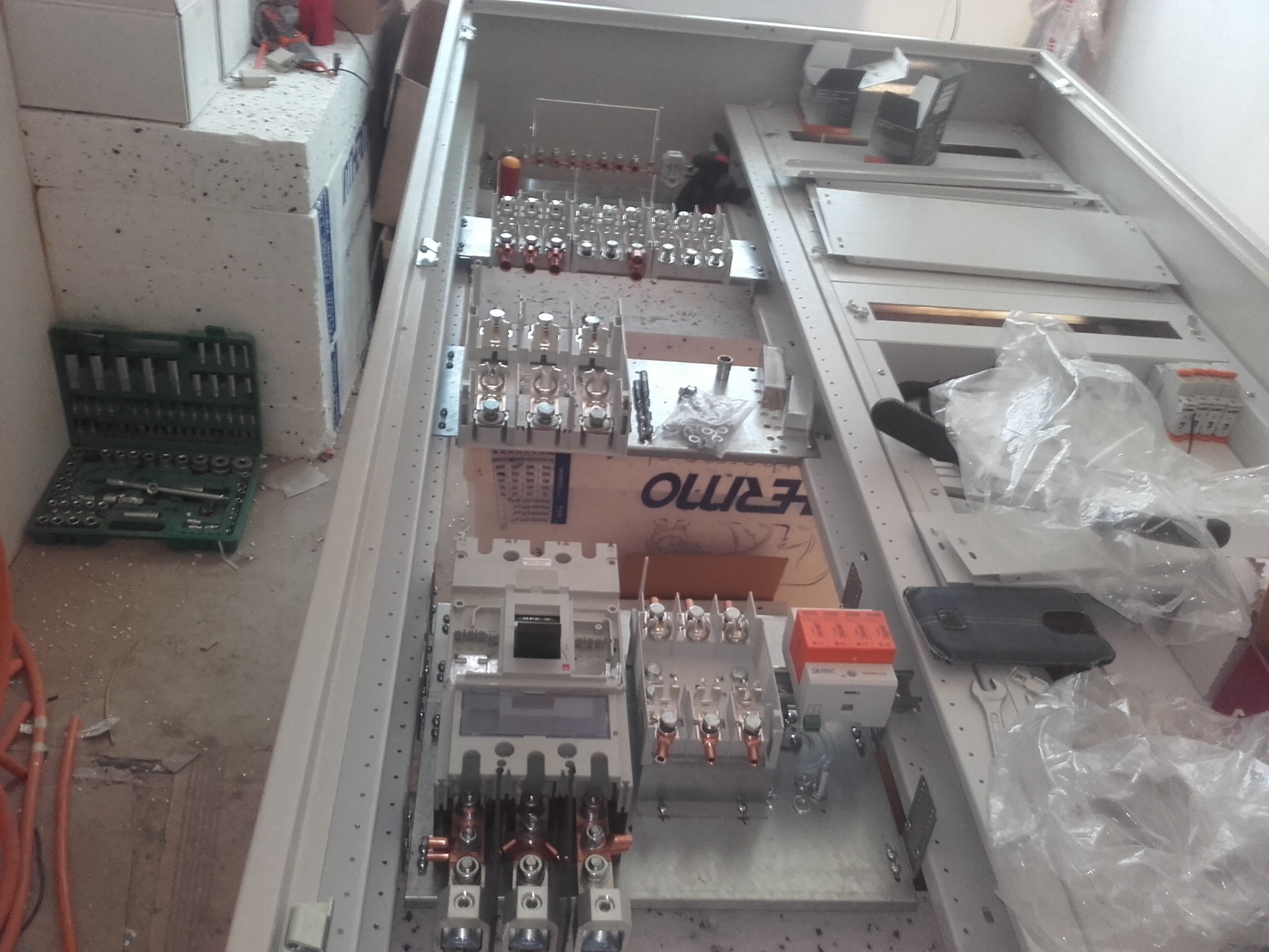 Wnętrze rozdzielnicy elektrycznej w trakcie montażu, widoczne liczne zabezpieczenia, styczniki, listwy zaciskowe, narzędzia i elementy montażowe, otwarte pudełko z kluczami nasadowymi po lewej...