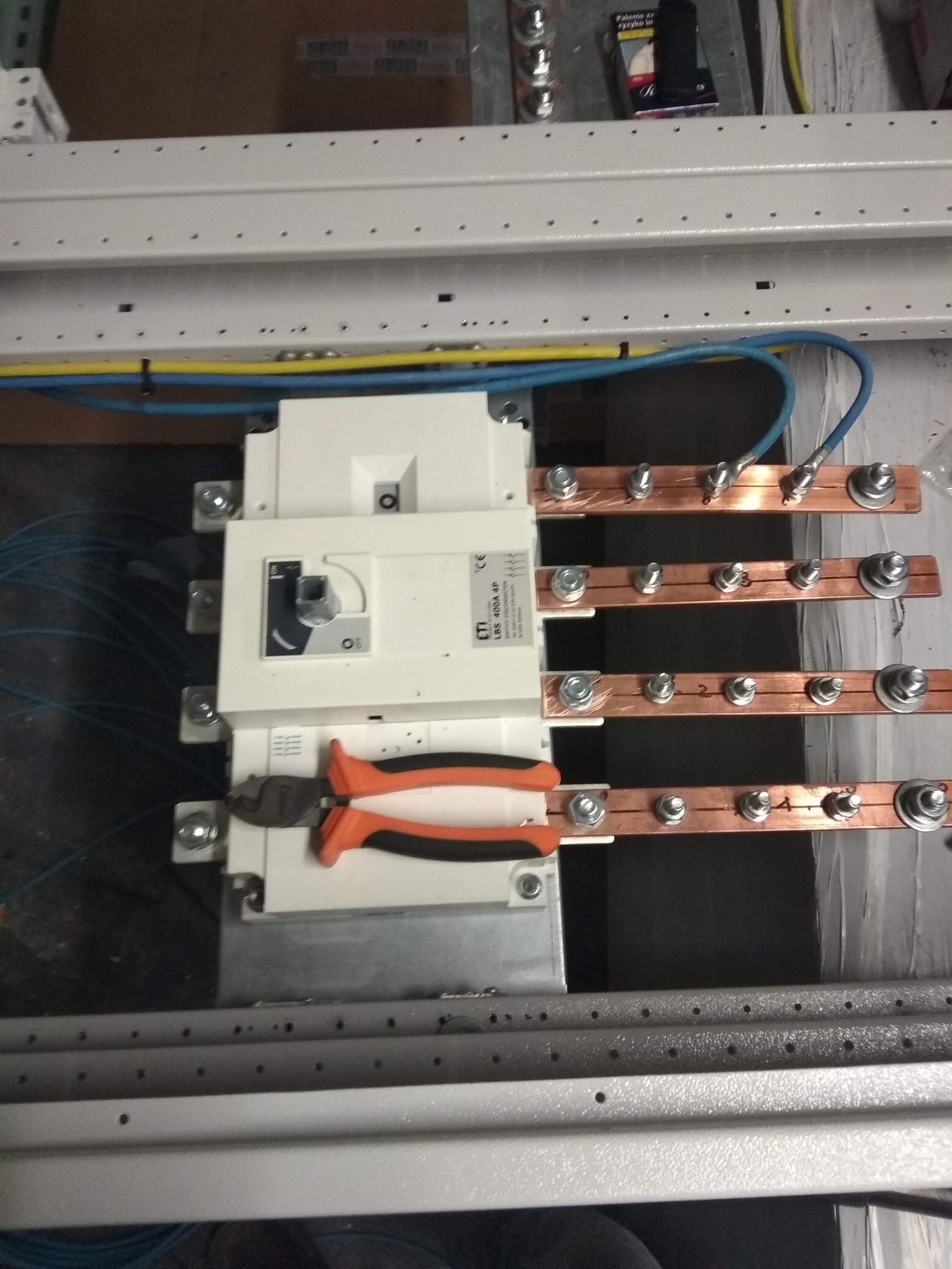 Rozdzielnica elektryczna z zamontowanym wyłącznikiem i szynami miedzianymi, widoczne narzędzia - obcinaczki z pomarańczowymi rękojeściami, przewody w kolorach niebieskim i żółtym.