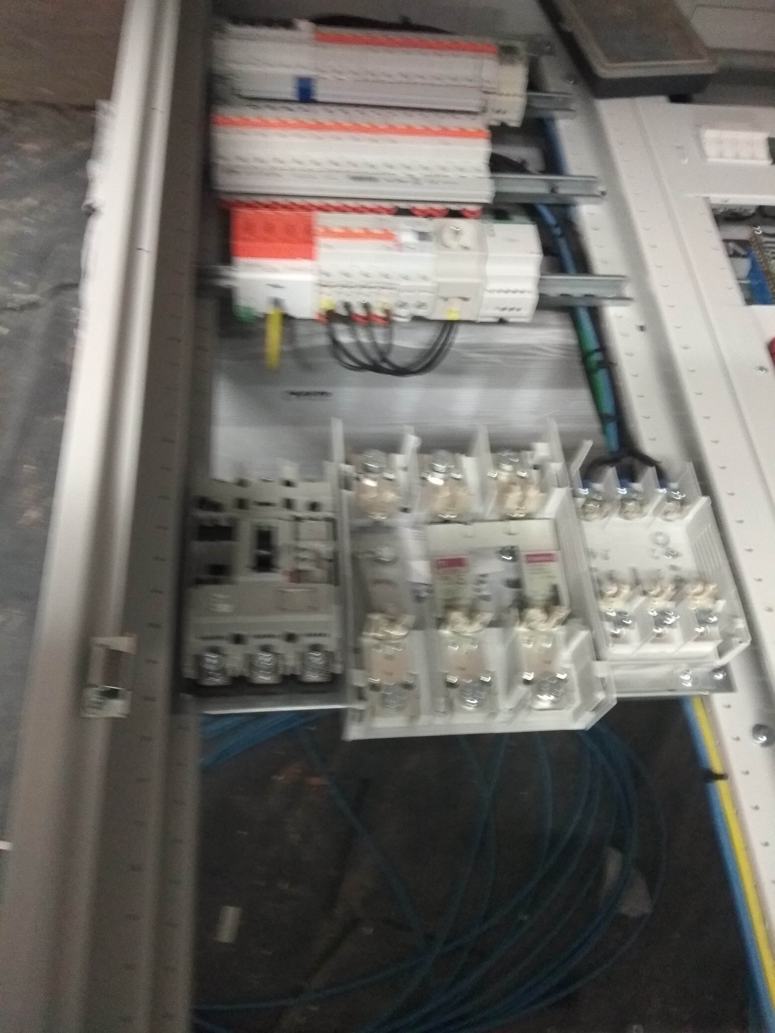 Wnętrze szafy rozdzielczej z zainstalowanymi bezpiecznikami, stycznikami i okablowaniem, widoczne przewody w kolorach niebieskim i żółto-zielonym, ułożone w dolnej części szafy.