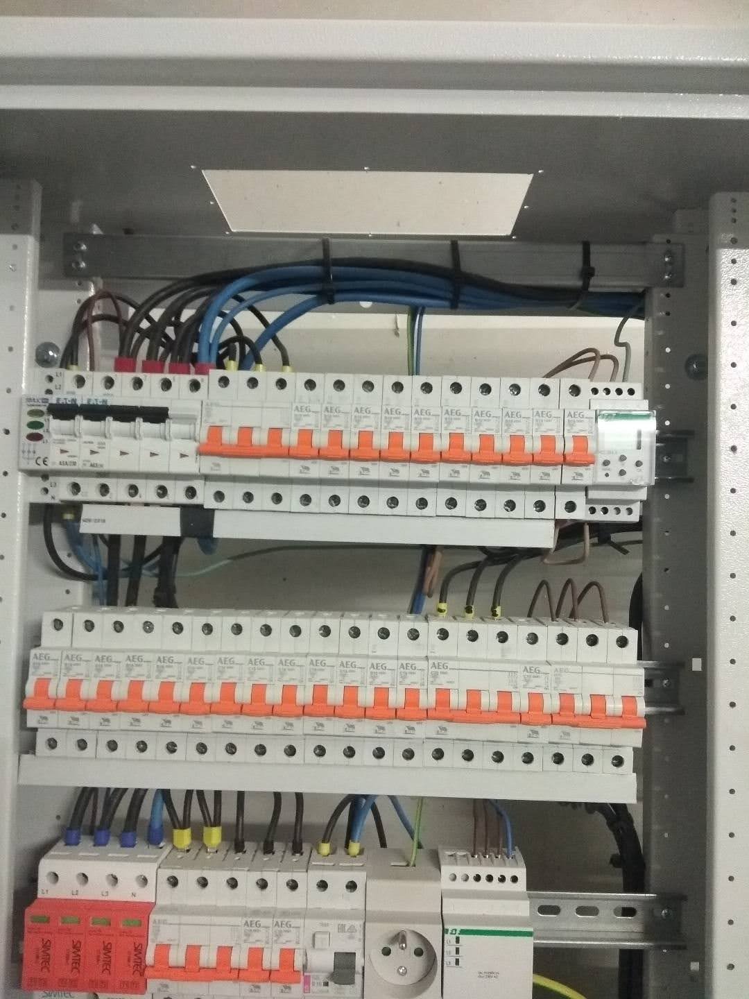 Rozdzielnica elektryczna z zainstalowanymi bezpiecznikami automatycznymi AEG, zabezpieczeniami przeciwprzepięciowymi Simtec i gniazdem elektrycznym, widoczne przewody w różnych kolorach.