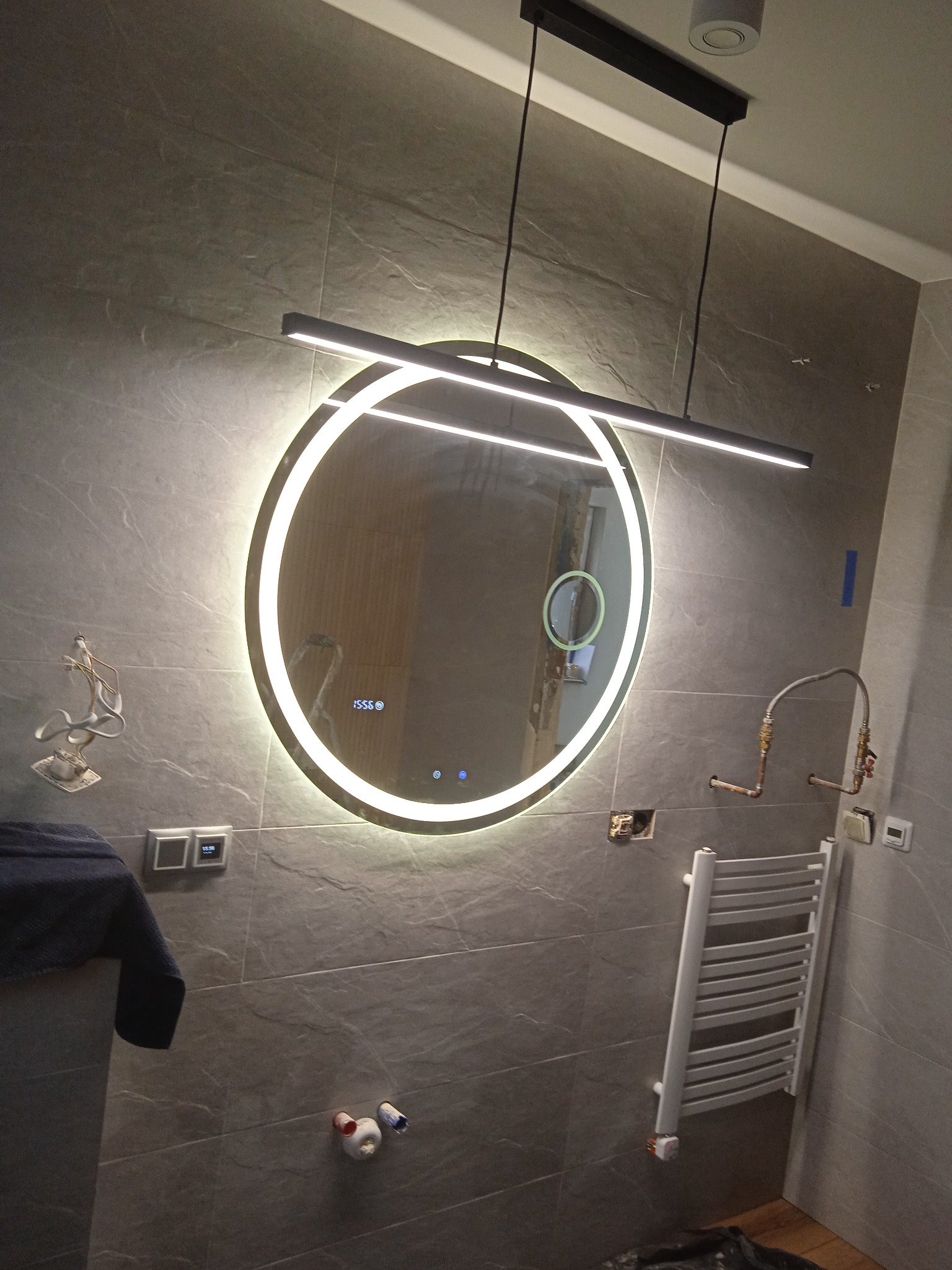 Nowoczesna łazienka w trakcie remontu z okrągłym lustrem LED, wiszącą lampą liniową, grzejnikiem drabinkowym i widocznymi instalacjami hydraulicznymi w ścianie wyłożonej szarymi płytkami...