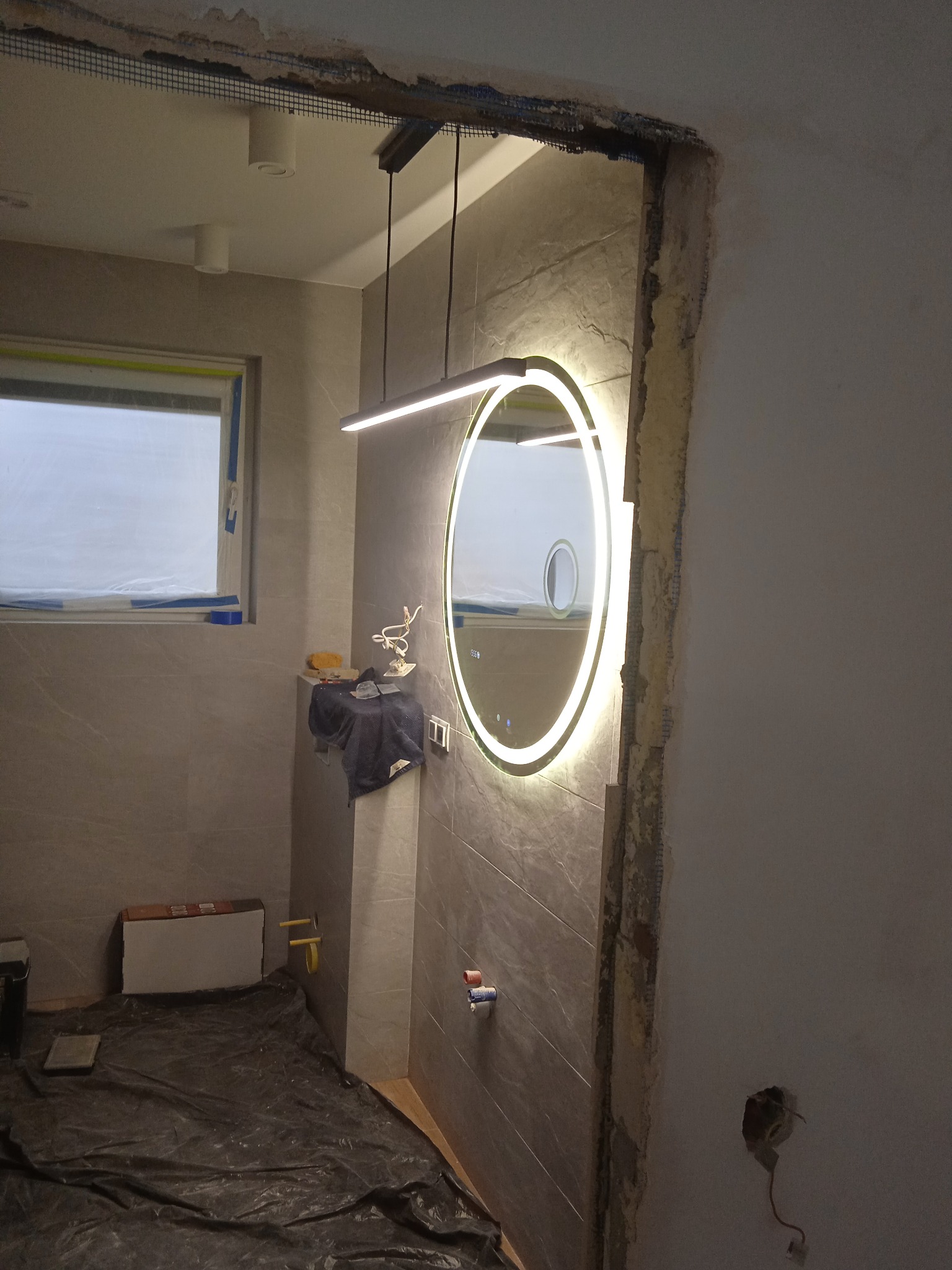 Łazienka w trakcie remontu: podwieszana lampa LED, okrągłe lustro z podświetleniem LED, widoczne przewody, zabezpieczona folią podłoga, ściany w trakcie wykończenia.