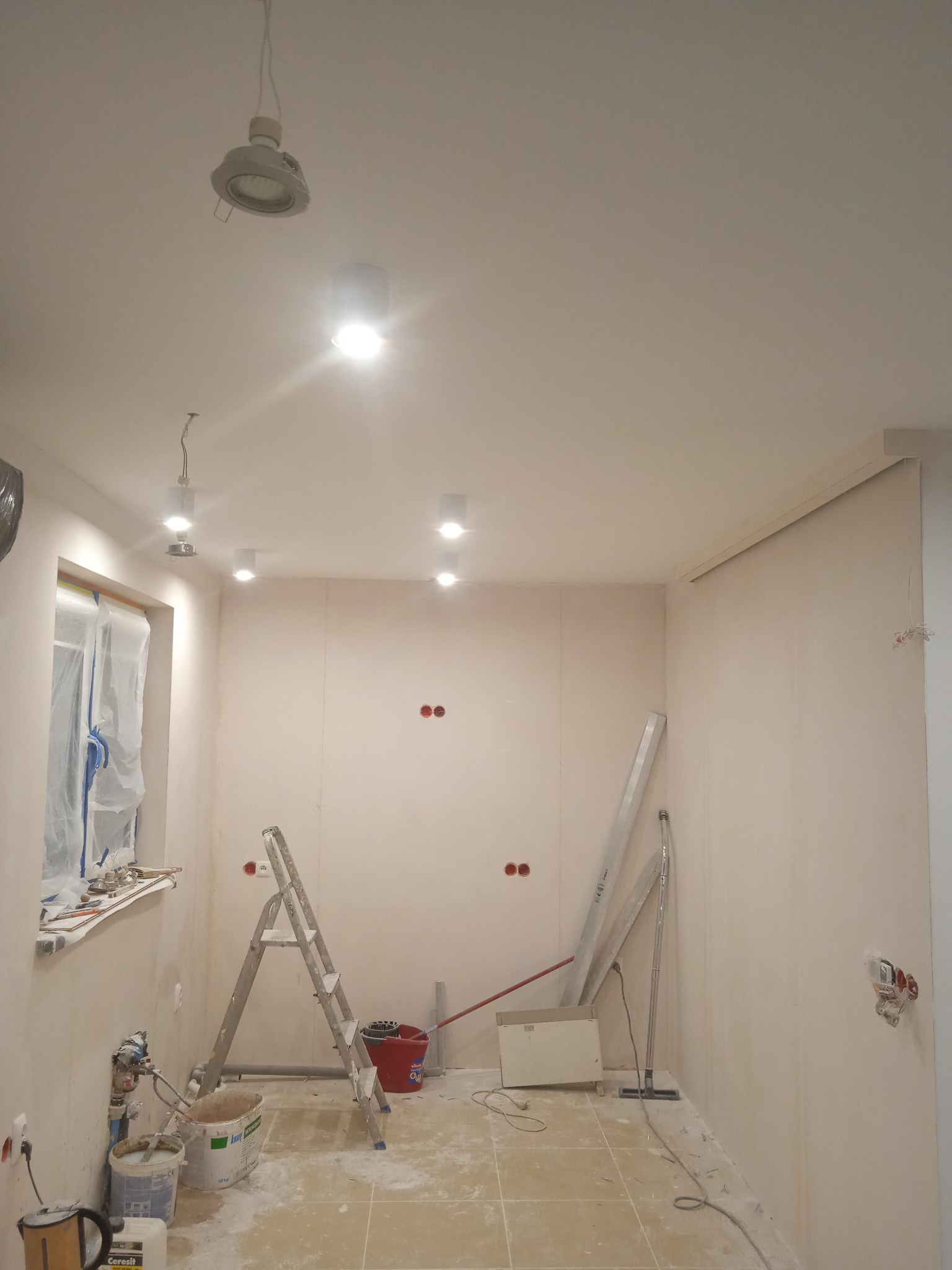 Wnętrze w trakcie remontu, widoczne puszki elektryczne w ścianie, drabina, narzędzia malarskie i przygotowane punkty świetlne na suficie.