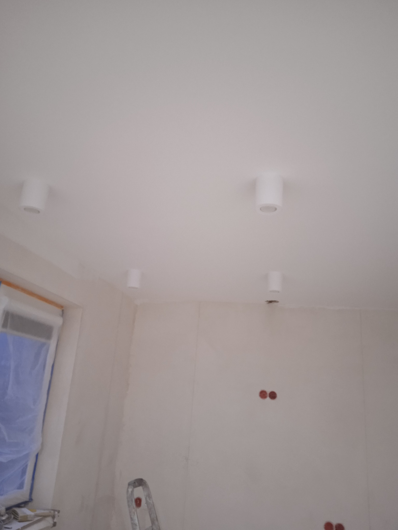 Białe, cylindryczne lampy sufitowe zamontowane w świeżo wykończonym pomieszczeniu, widoczne ślady szpachlowania na ścianach, okno zabezpieczone folią malarską, fragment drabiny w dolnej części kadru.