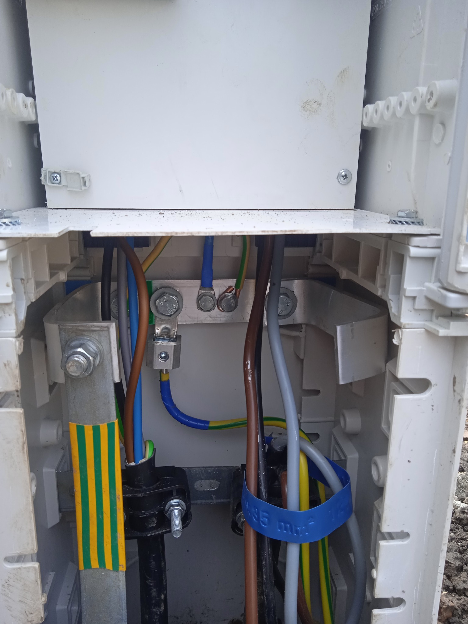 Wnętrze skrzynki elektrycznej z widocznymi przewodami o różnych kolorach (brązowy, niebieski, żółto-zielony, szary), złączkami śrubowymi i elementami mocującymi, w tym niebieską opaską z napisem...