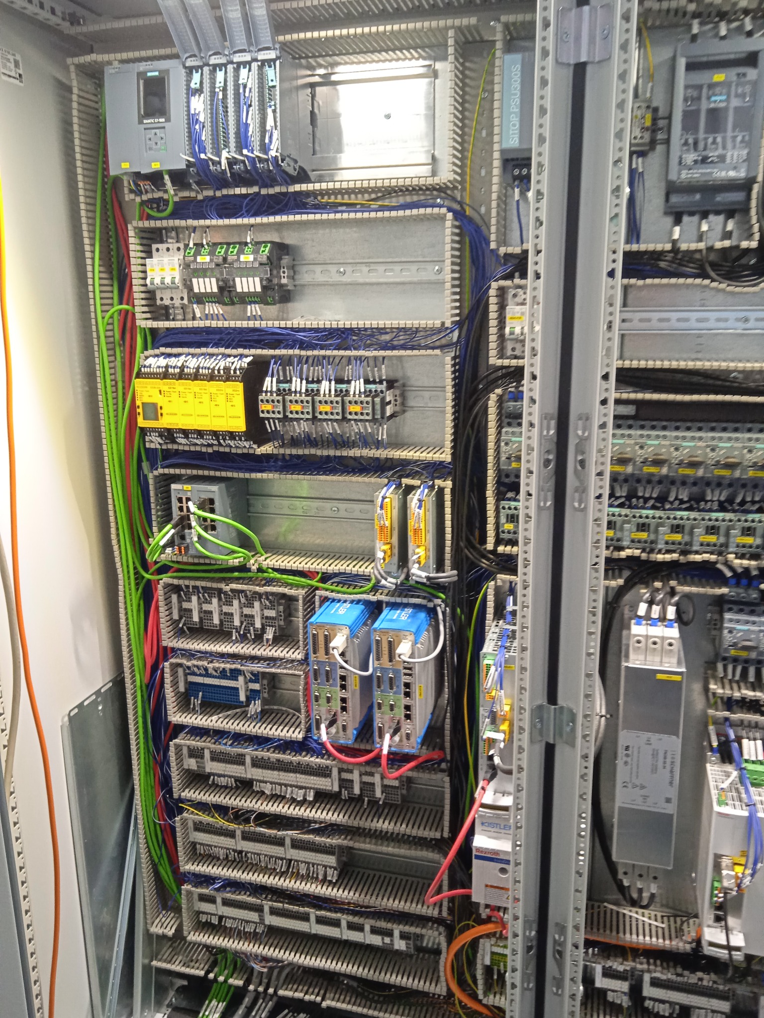 Rozdzielnica elektryczna z licznymi modułami sterującymi, przewodami w różnych kolorach (zielony, niebieski, żółty, czerwony), szynami montażowymi i elementami zabezpieczającymi. Widoczne...