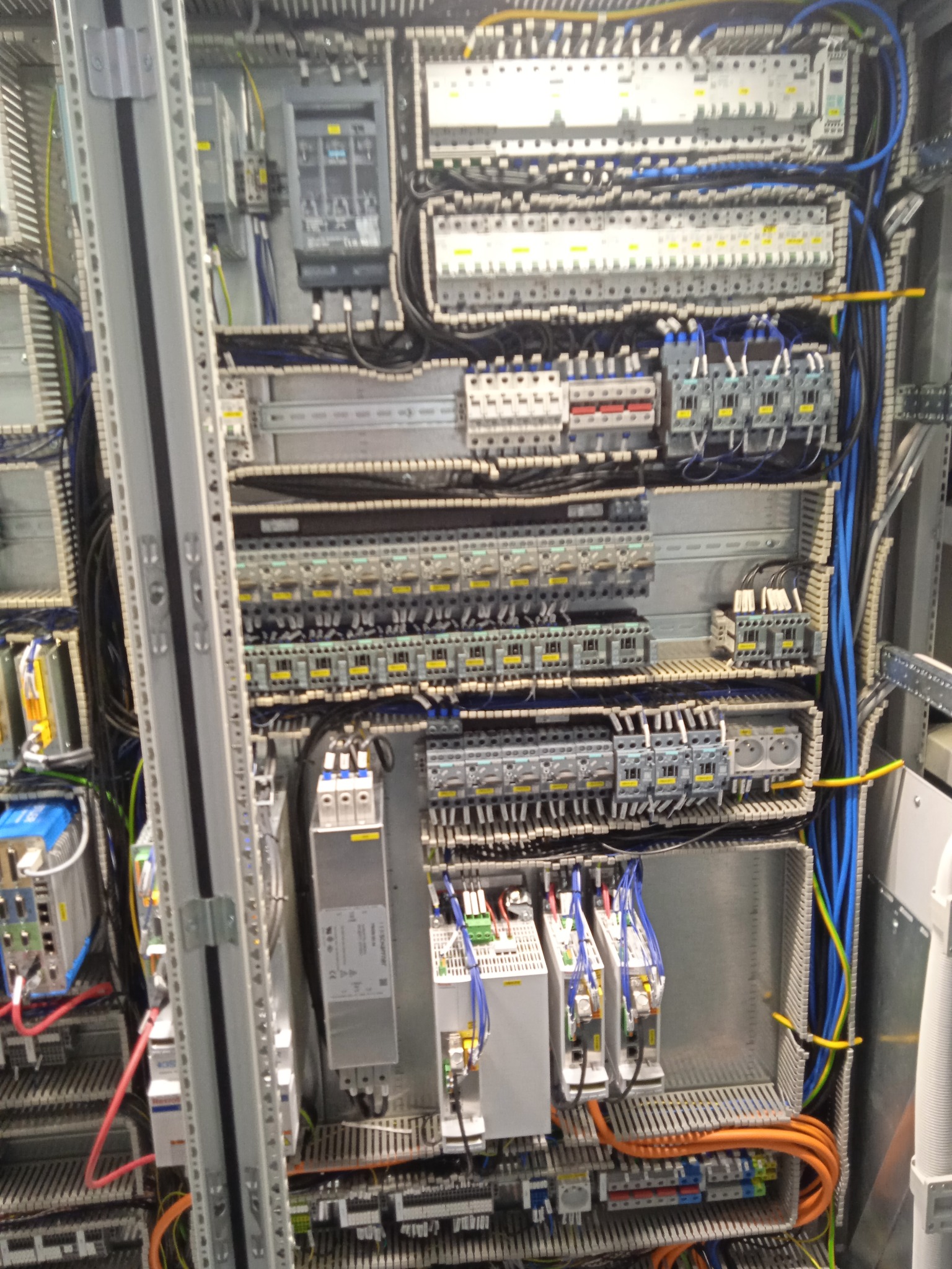 Rozdzielnica elektryczna z licznymi zabezpieczeniami, przewodami w różnych kolorach i modułami sterującymi, widoczne szyny montażowe i elementy mocujące.