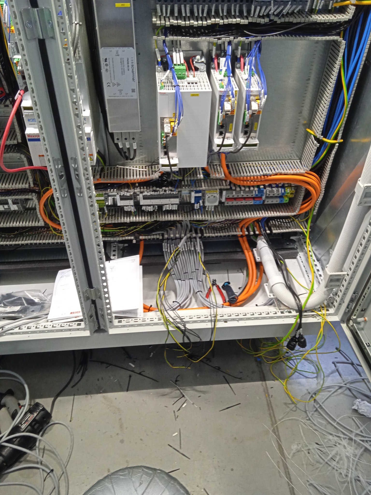 Wnętrze szafy sterowniczej z widocznymi elementami aparatury, przewodami w peszlach i luźnymi przewodami na podłodze, dokumentacja techniczna na półce.