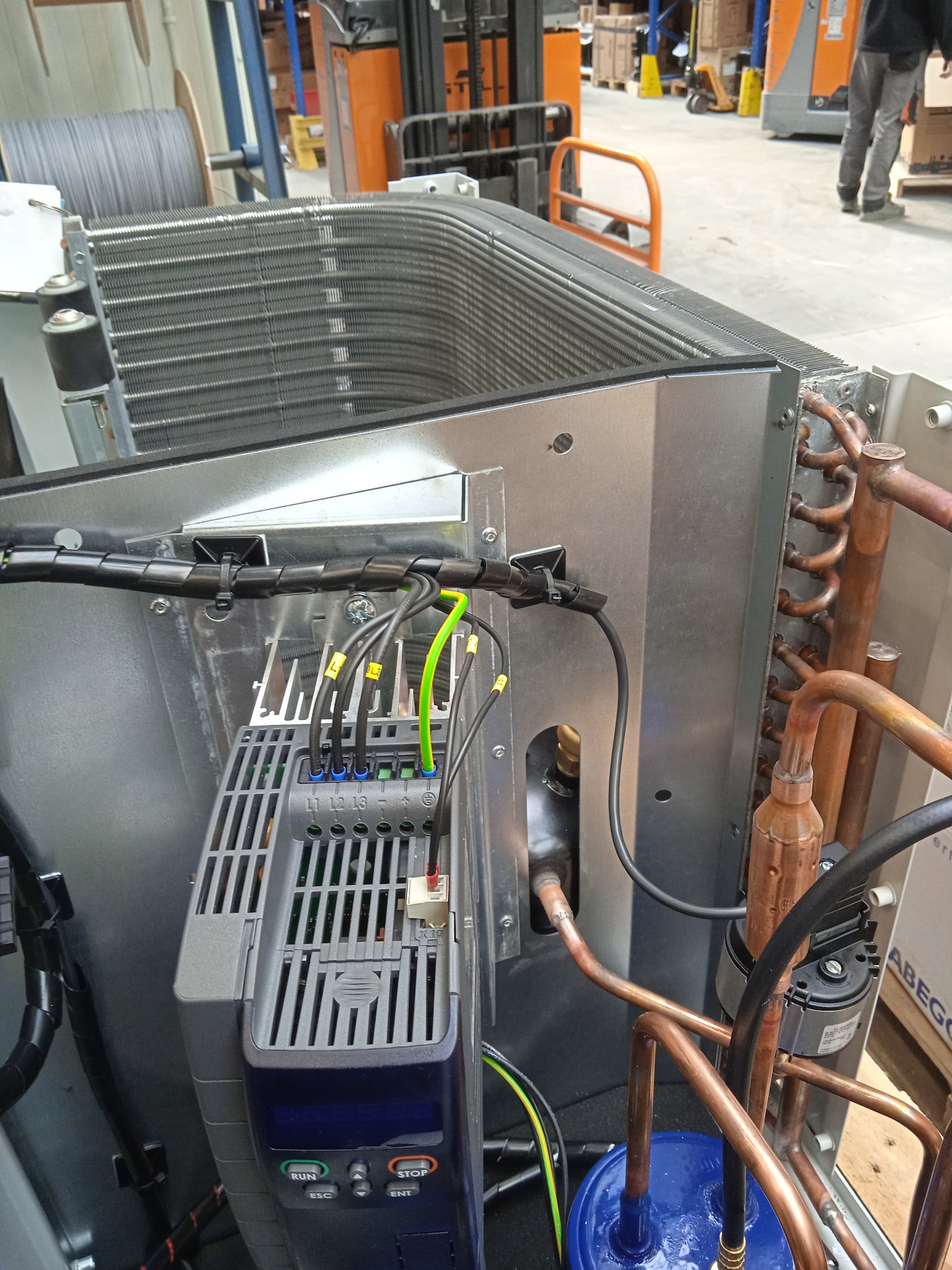 Widok z bliska na podłączenie elektryczne do klimatyzatora przemysłowego z widocznym falownikiem, przewodami oznaczonymi numerami, miedzianymi rurkami i elementami chłodzącymi, na tle hali...