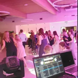 Sprzęt DJski z laptopem i oświetleniem scenicznym na tle tańczących osób w sali oświetlonej różowym światłem.