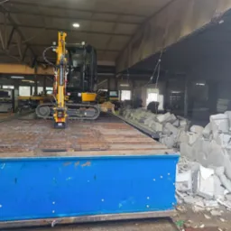 Żółta mini koparka gąsienicowa na platformie w hali, obok sterta gruzu po rozbiórce ściany, widoczny fragment zniszczonej konstrukcji budynku.