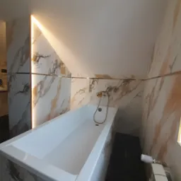 Wykończona łazienka na poddaszu ze skośnym sufitem, z wanną obudowaną płytkami imitującymi marmur, złotą armaturą i podświetleniem LED wzdłuż ściany.