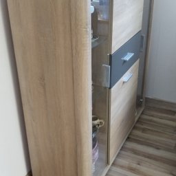 Złota raczka - Nowoczesna komoda z przeszklonymi półkami, na blacie ramki na zdjęcia, figurka anioła i inne dekoracje, ustawiona na panelach podłogowych.