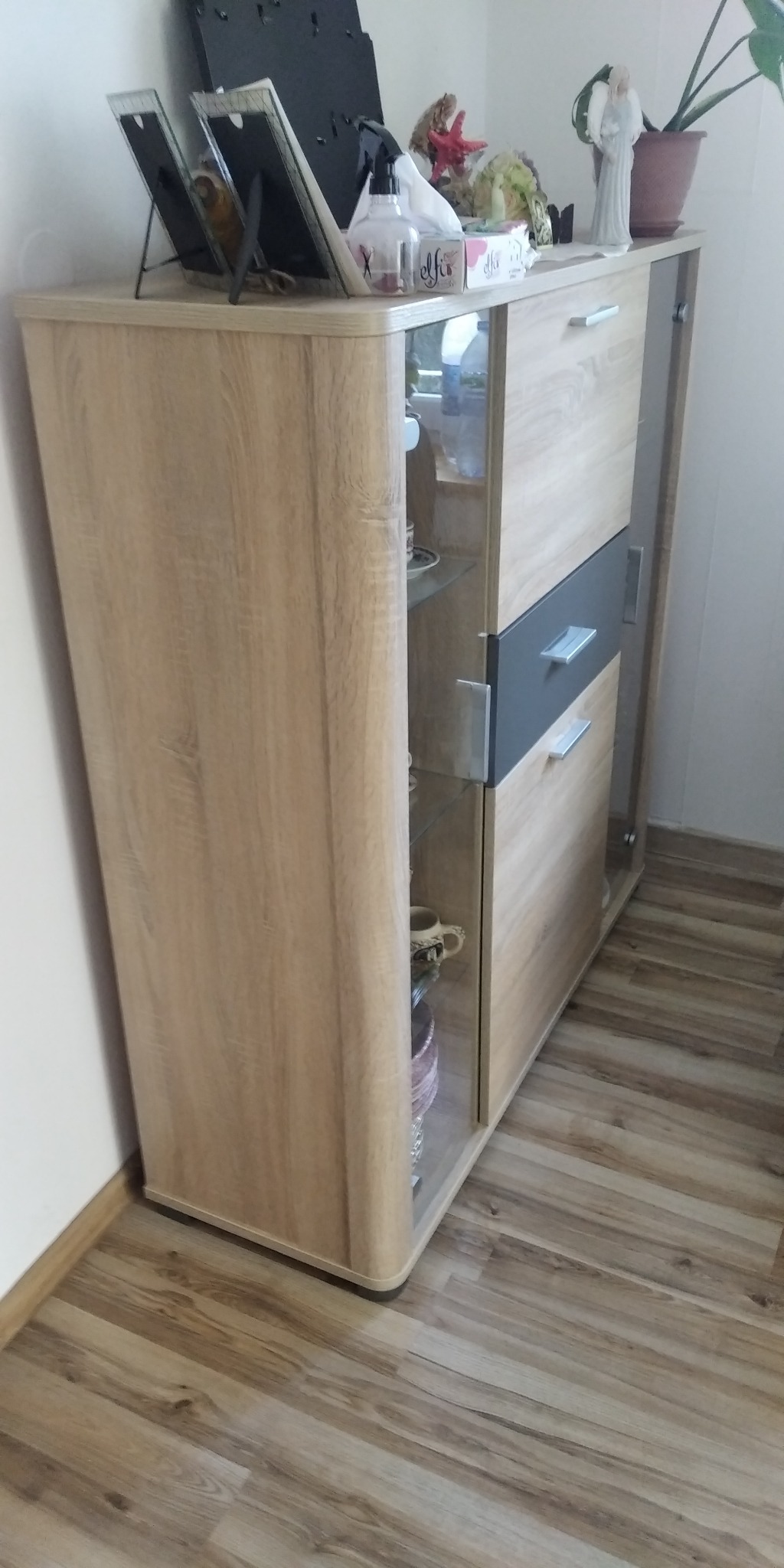 Nowoczesna komoda z przeszklonymi półkami, na blacie ramki na zdjęcia, figurka anioła i inne dekoracje, ustawiona na panelach podłogowych.