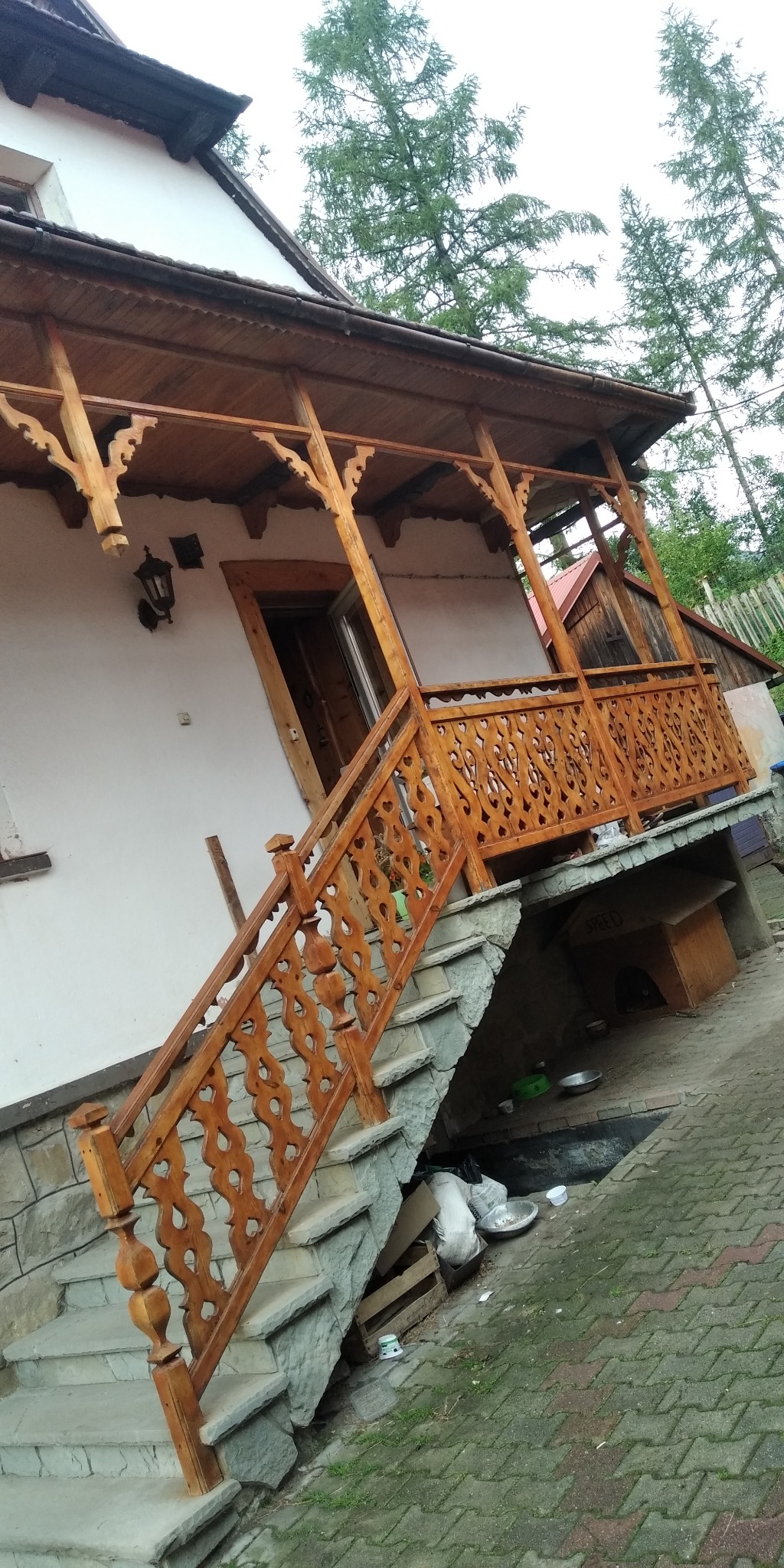 Drewniany, zdobiony ganek z balustradą w stylu regionalnym, prowadzący do wejścia domu w Rokicinach Podhalańskich. Schody wyłożone kamieniem, widoczny fragment podwórka.