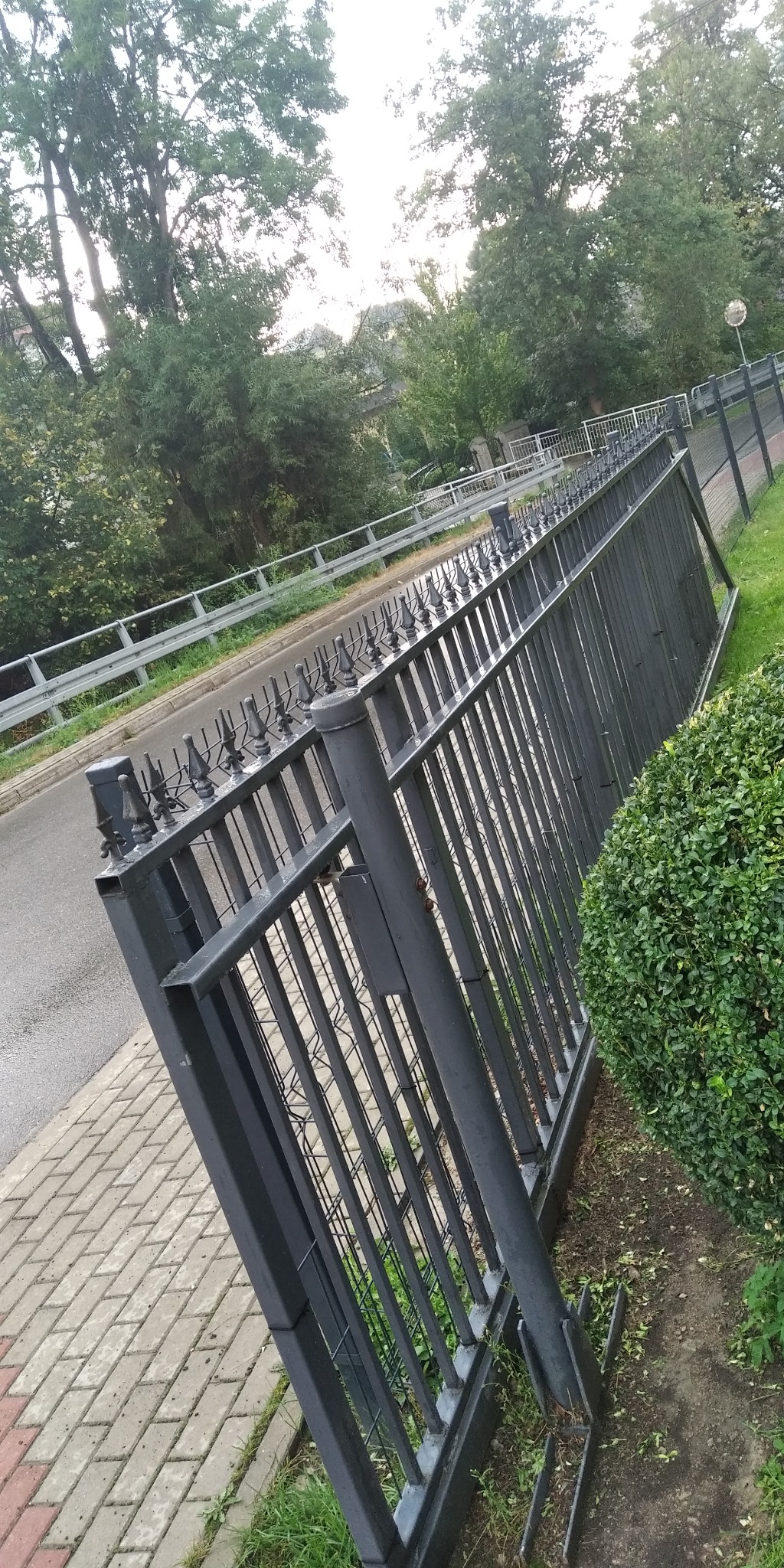 Metalowe ogrodzenie z ozdobnymi grotami, oddzielające chodnik z kostki brukowej od jezdni, z zielenią w tle.