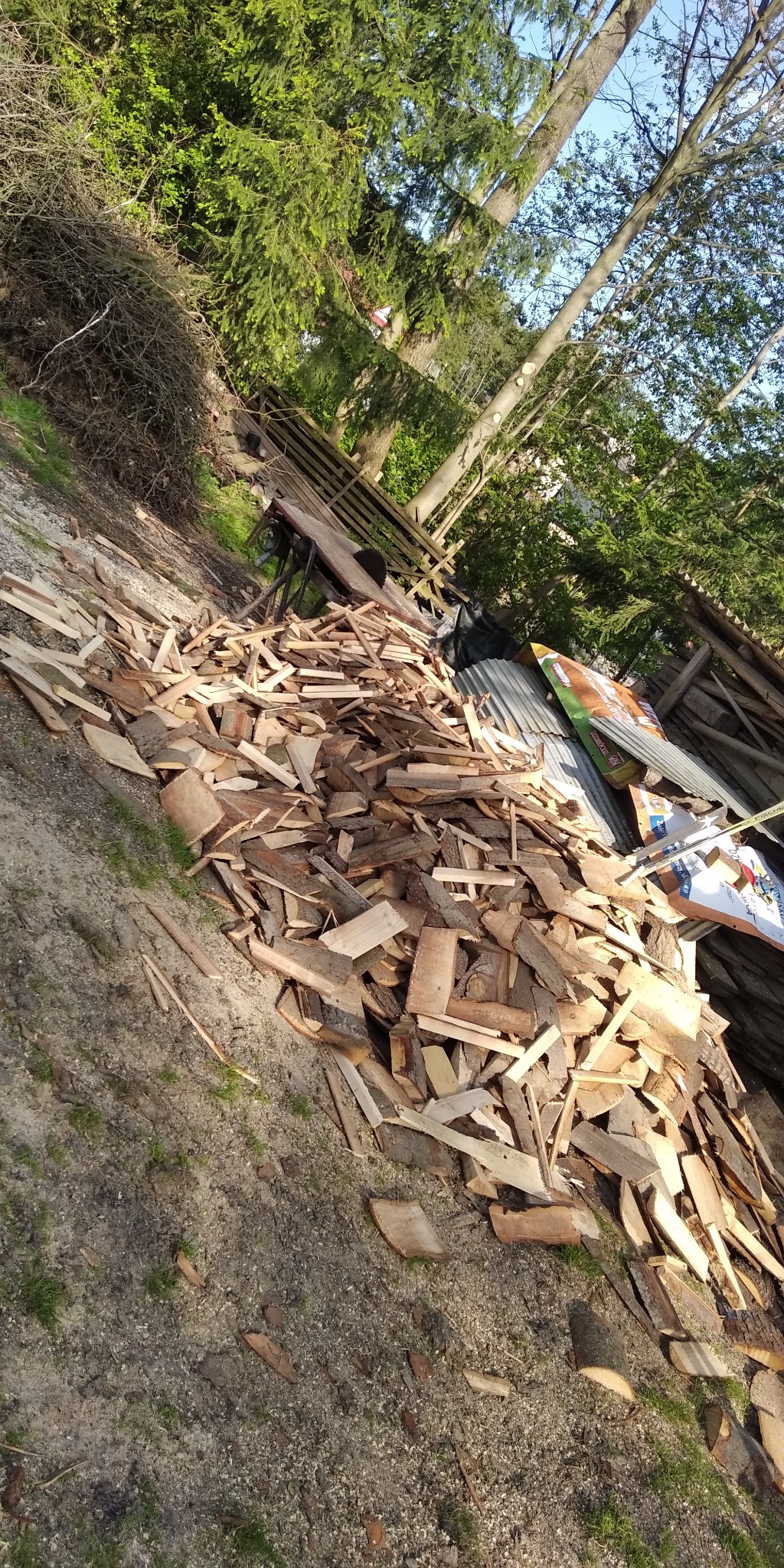 Stos pociętego drewna leżący na ziemi, w tle widać drzewa i fragment konstrukcji drewnianej.