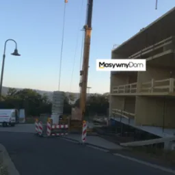 Budowa domu modułowego: dźwig podnosi prefabrykowane elementy konstrukcyjne, widoczny szkielet budynku, rusztowania, oraz zaparkowany bus firmy budowlanej z widocznym logo.