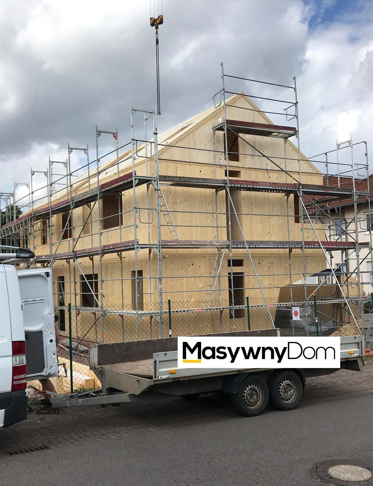 Wznoszenie dwukondygnacyjnego domu z drewnianych paneli, z widocznym rusztowaniem, przyczepą i dźwigiem w pochmurny dzień.