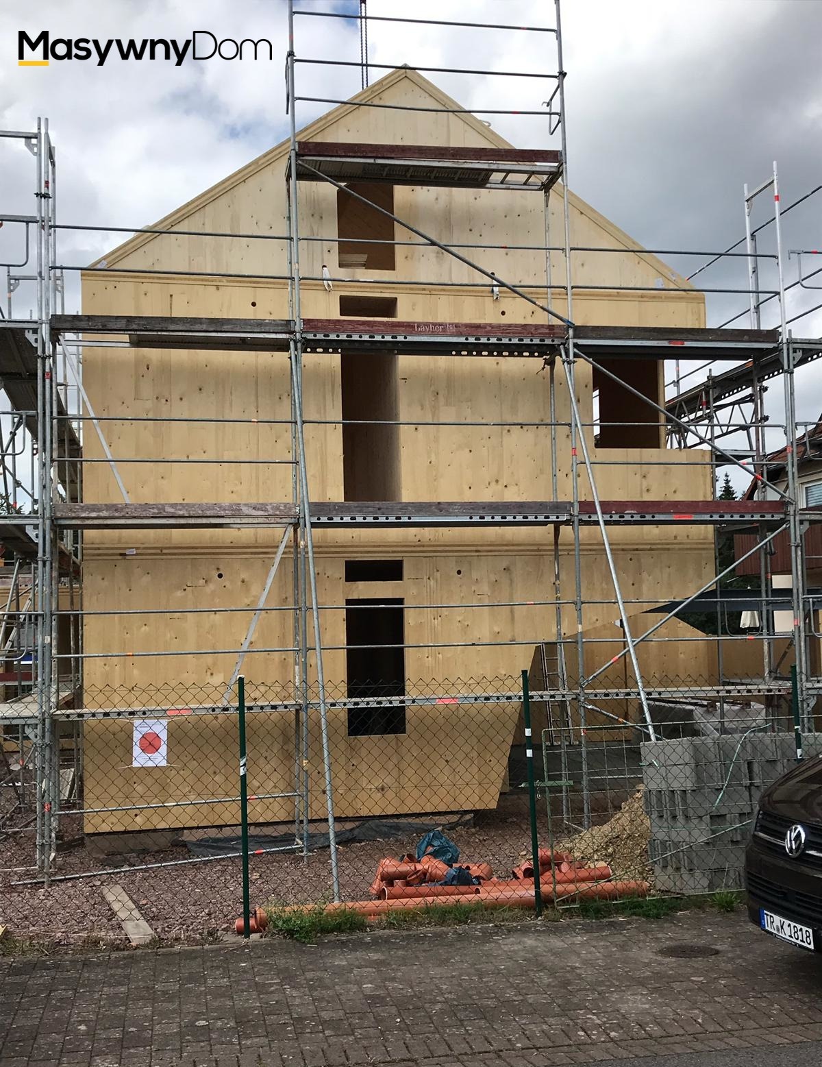 Wznoszona konstrukcja domu z drewnianych paneli na placu budowy, widoczne rusztowania, ogrodzenie z siatki i elementy instalacji kanalizacyjnej.