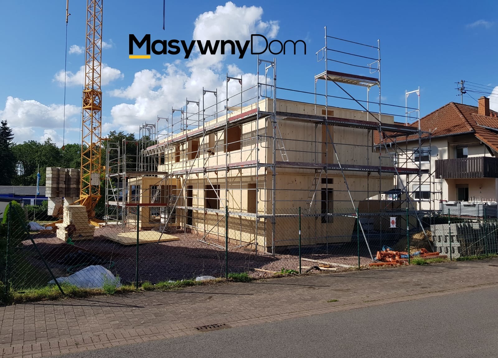 Dwukondygnacyjny dom w trakcie budowy, widoczne drewniane panele szkieletowe, rusztowania, żuraw budowlany w tle, logo firmy na zdjęciu.