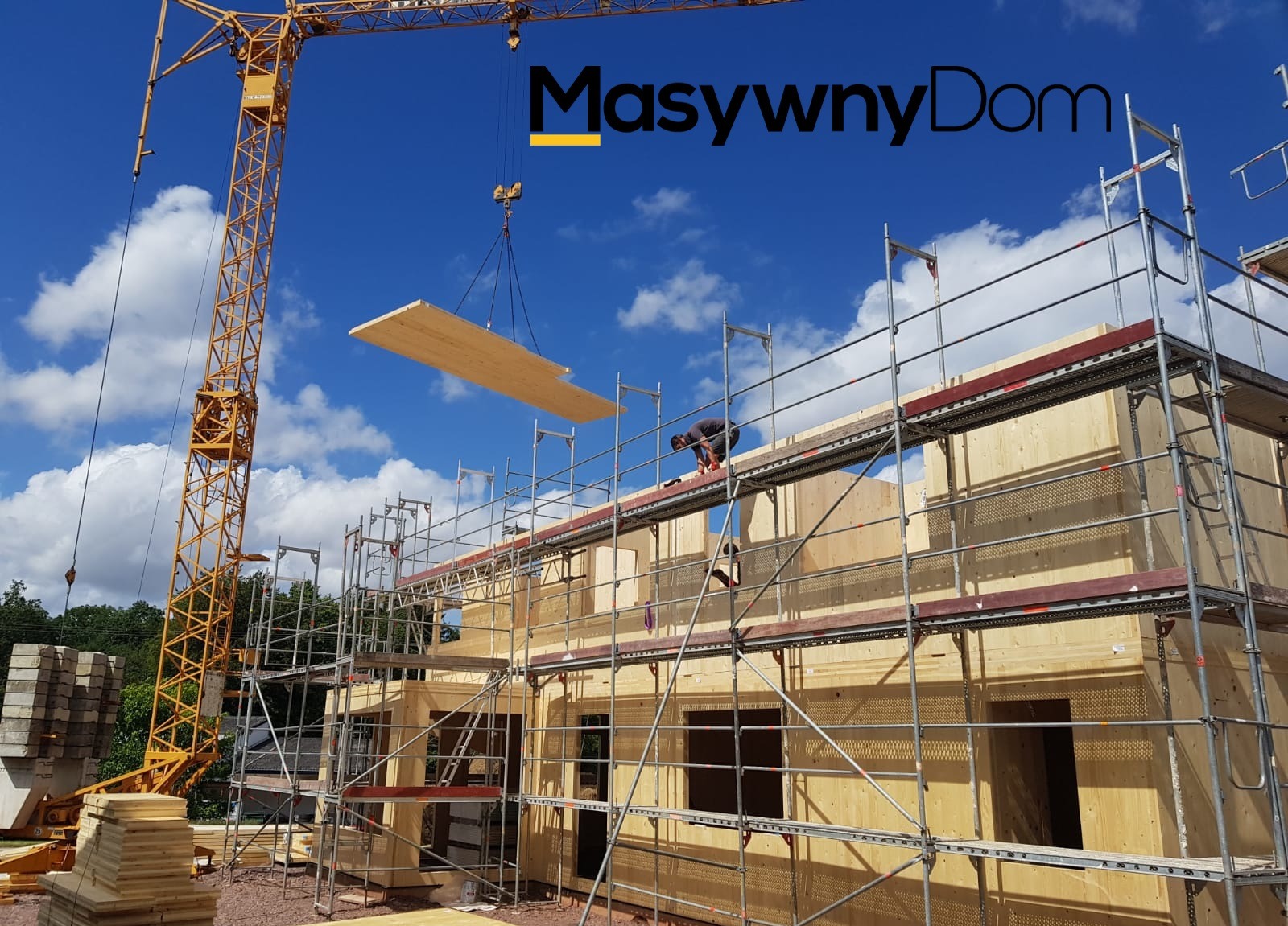 Montaż drewnianej konstrukcji domu na placu budowy z użyciem dźwigu, widoczne rusztowania i pracownik na górze.