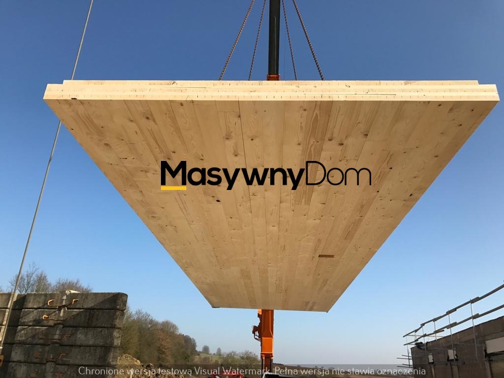 Panel drewniany z widocznym logo firmy 'MasywnyDom' podnoszony dźwigiem na tle błękitnego nieba, z widocznymi elementami konstrukcji oraz stertą betonowych bloków w dolnej części kadru.