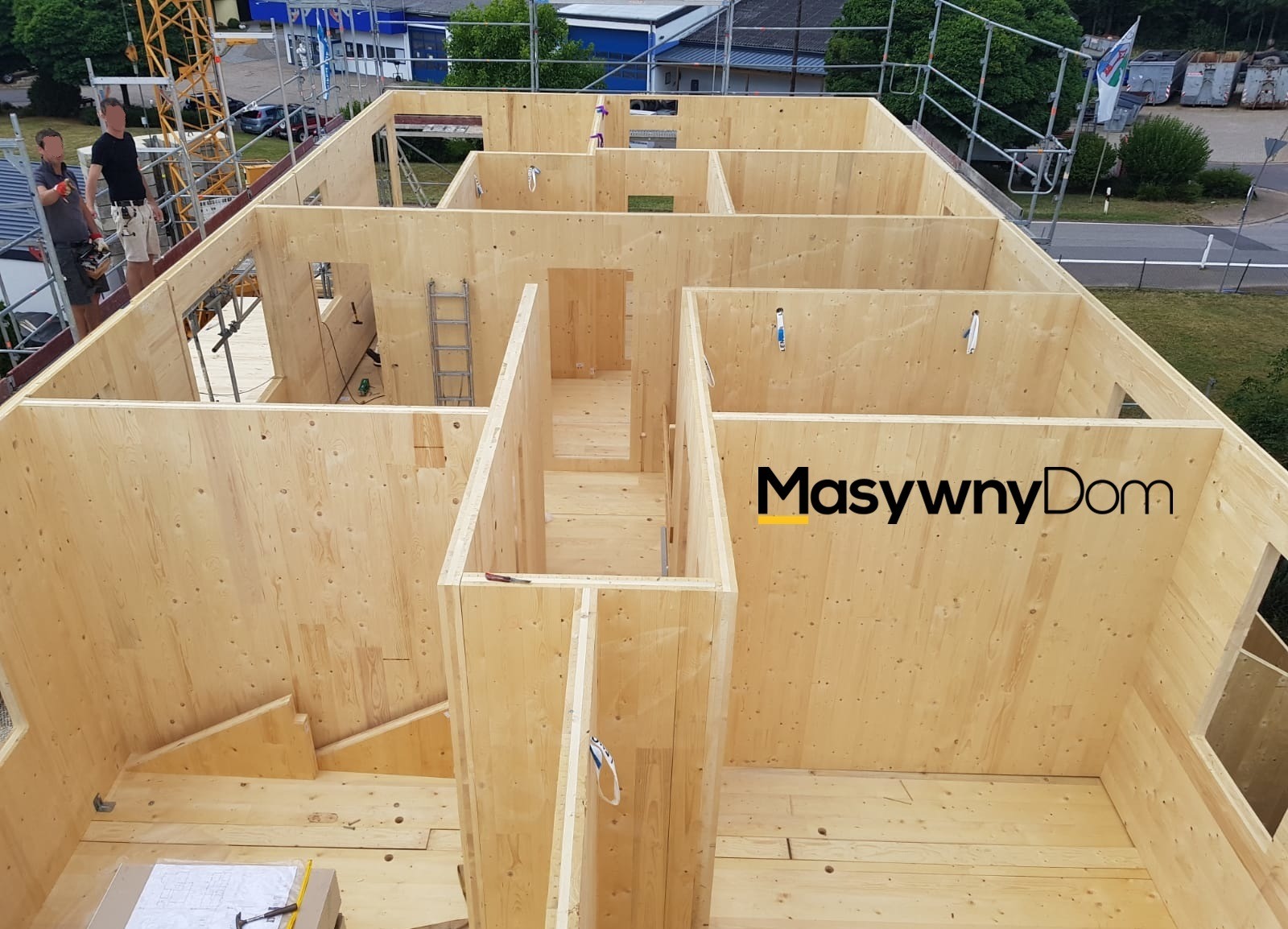 Wnętrze w trakcie budowy domu z drewnianych paneli, widoczne ściany działowe, strop, drabina, narzędzia i pracownicy na rusztowaniu, logo firmy na jednej ze ścian.