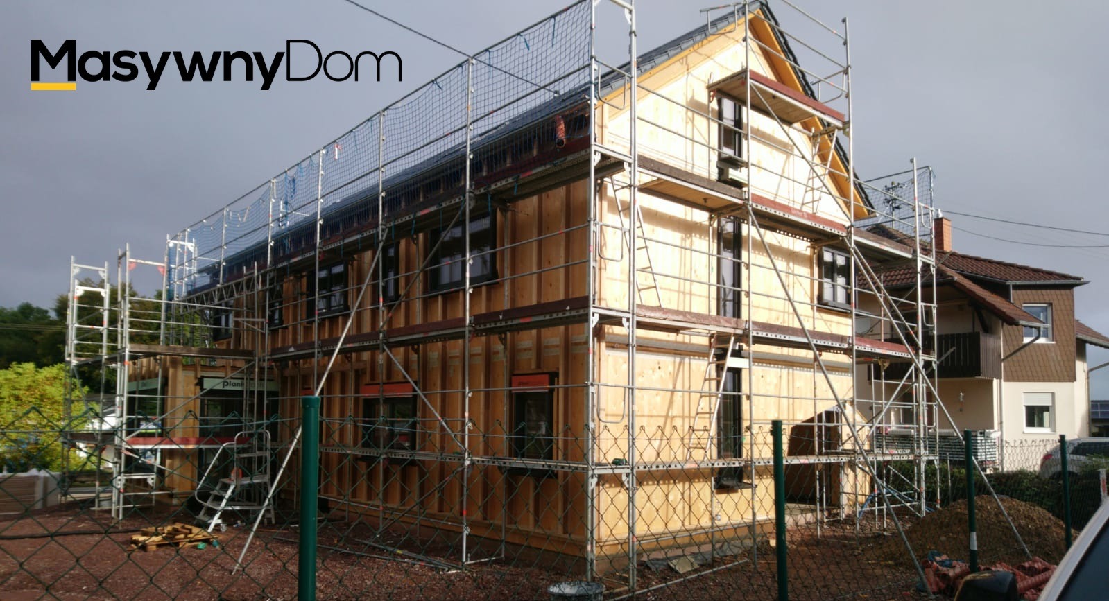 Dom w trakcie budowy, obłożony drewnianymi panelami, otoczony rusztowaniem i siatką zabezpieczającą, widoczny napis 'MasywnyDom' w lewym górnym rogu.