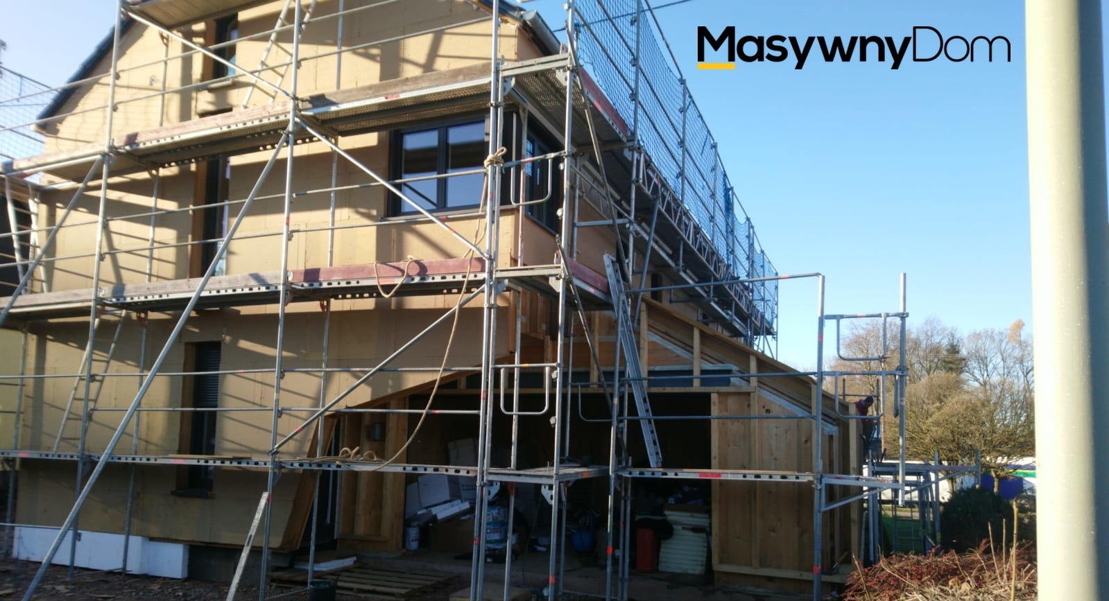 Wznoszony dom w konstrukcji szkieletowej, z widocznym rusztowaniem i częściowo ocieplonymi ścianami zewnętrznymi, logo firmy w prawym górnym rogu.