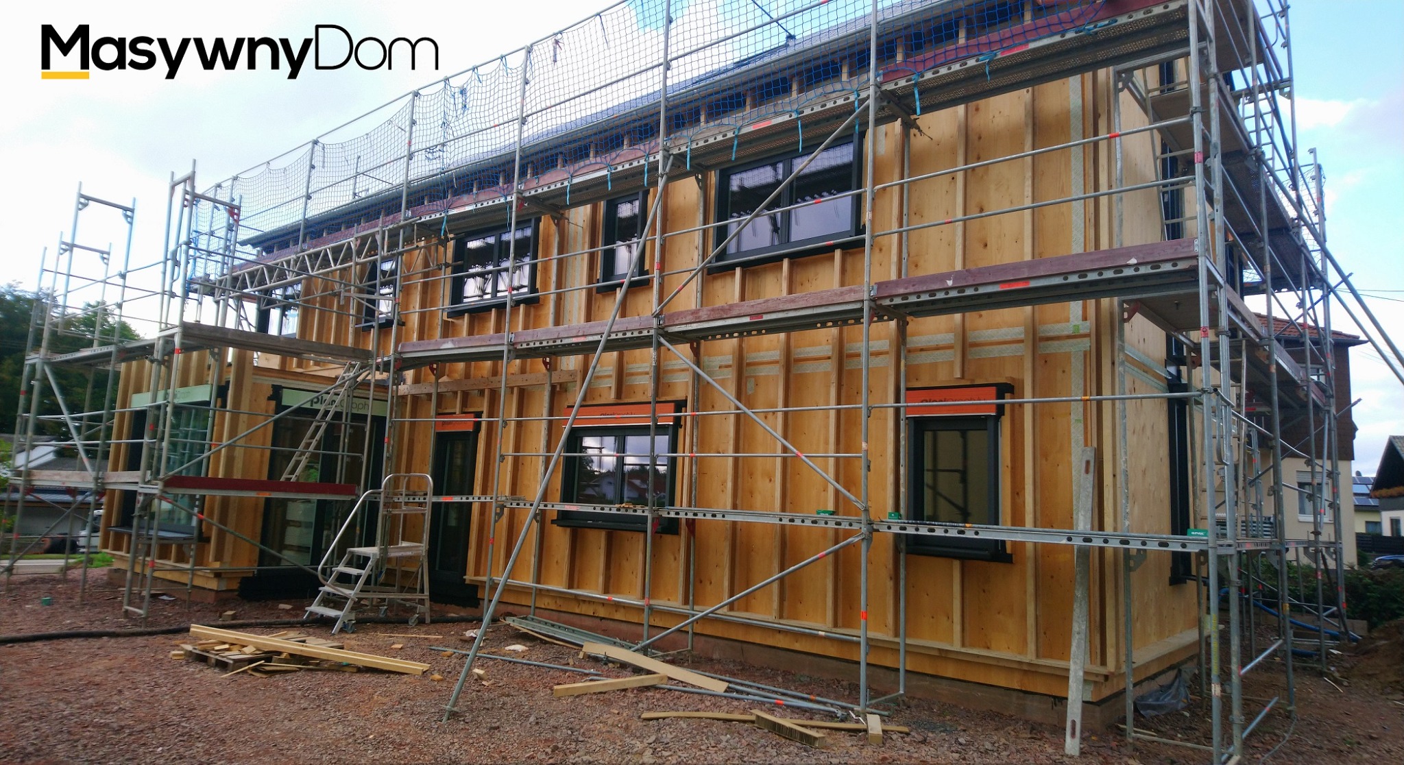 Wznoszenie dwukondygnacyjnego domu szkieletowego z widoczną konstrukcją drewnianą i rusztowaniami, częściowo ocieplonego, z czarnymi oknami.