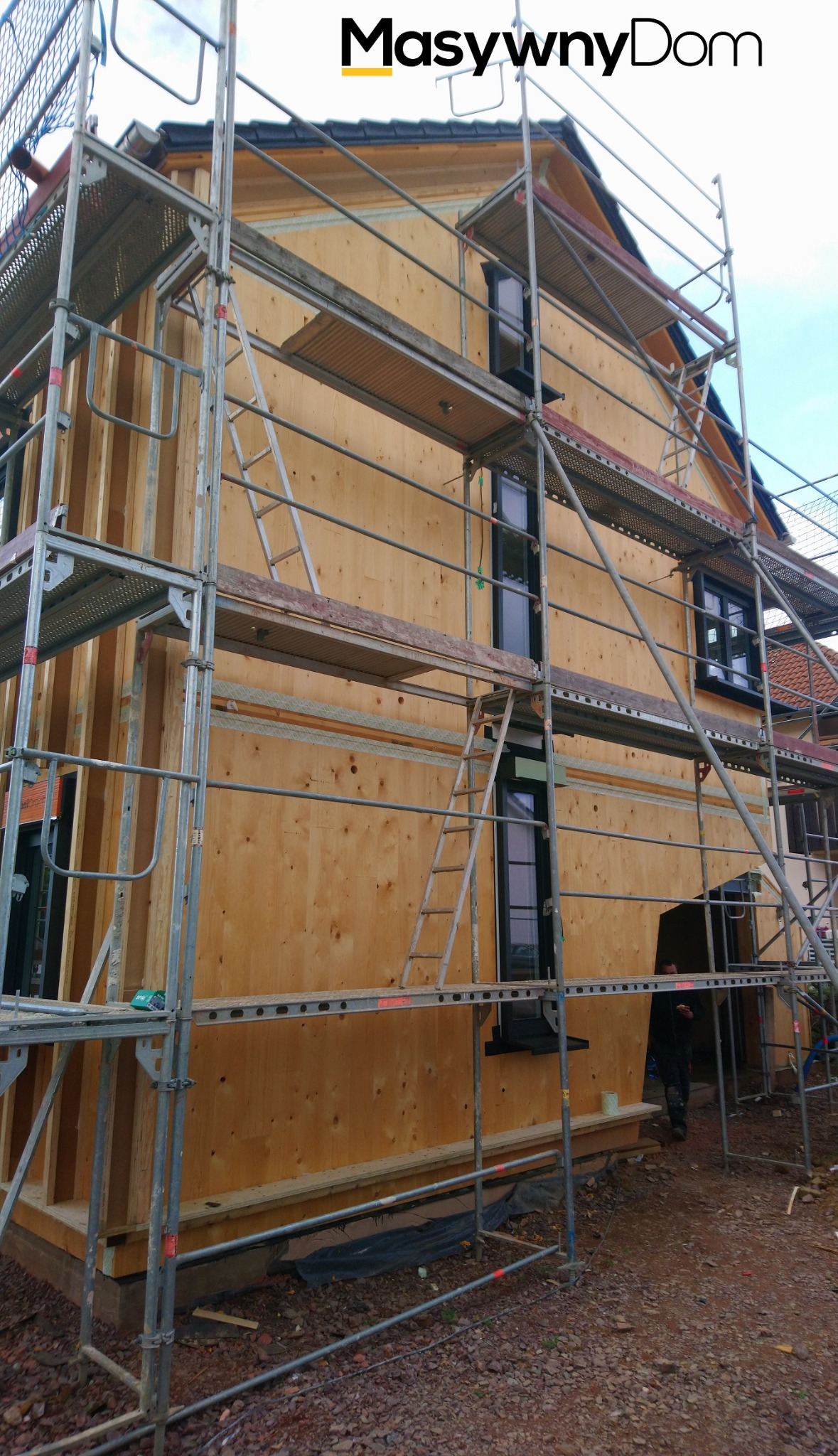 Surowa elewacja domu w trakcie budowy, obłożona drewnianymi panelami, widoczna konstrukcja rusztowania i czarne ramy okienne.