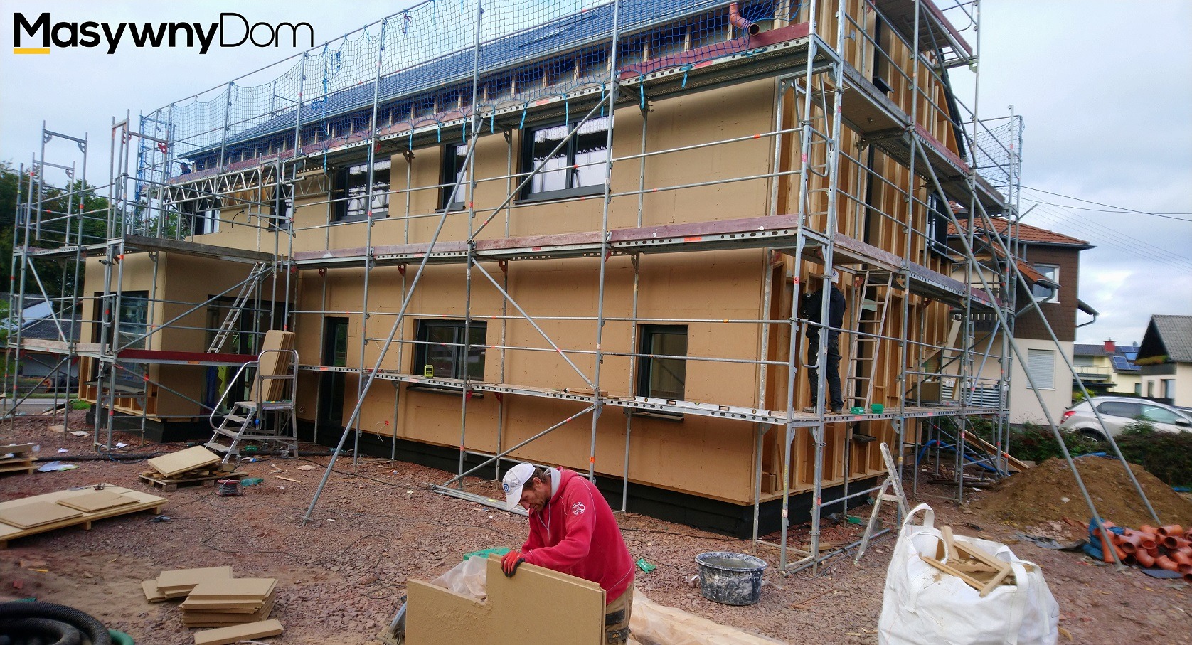 Dwukondygnacyjny budynek w trakcie montażu, otoczony rusztowaniem, z widocznymi elementami konstrukcyjnymi i pracownikiem w czerwonej bluzie przygotowującym panele elewacyjne na placu budowy.