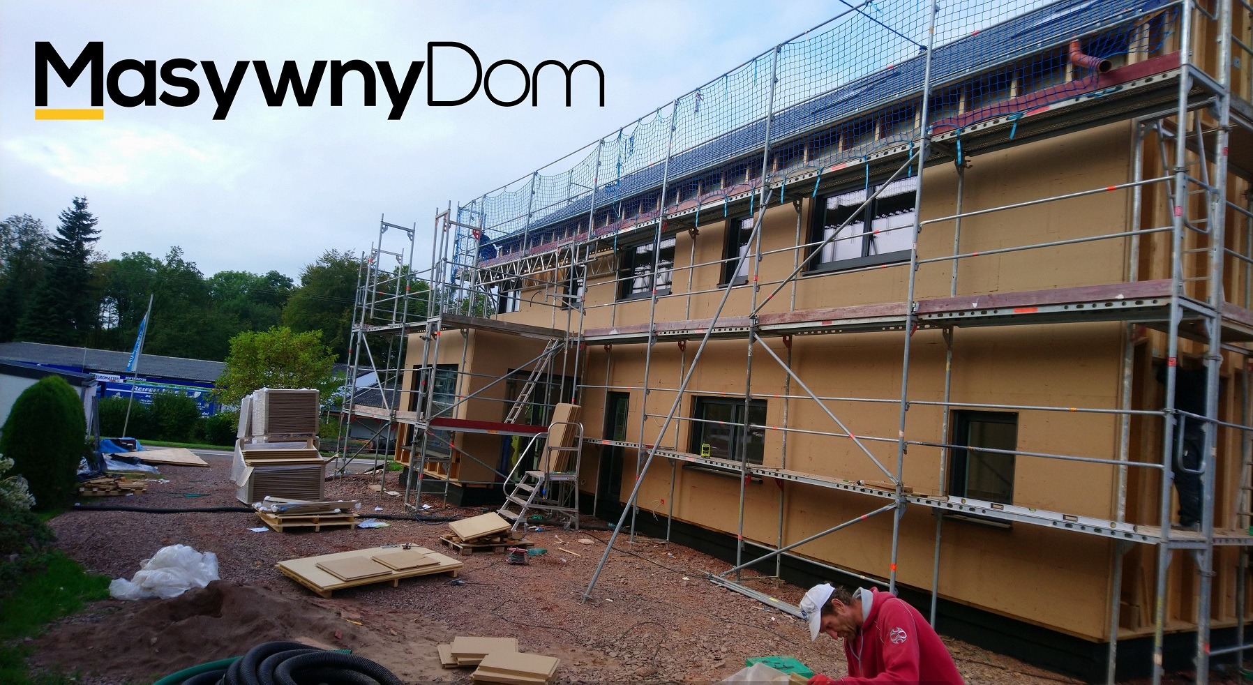 Wznoszenie dwukondygnacyjnego budynku z drewnianą konstrukcją szkieletową, osłoniętego siatką zabezpieczającą i rusztowaniami, z widocznym pracownikiem w czerwonej bluzie na pierwszym planie.