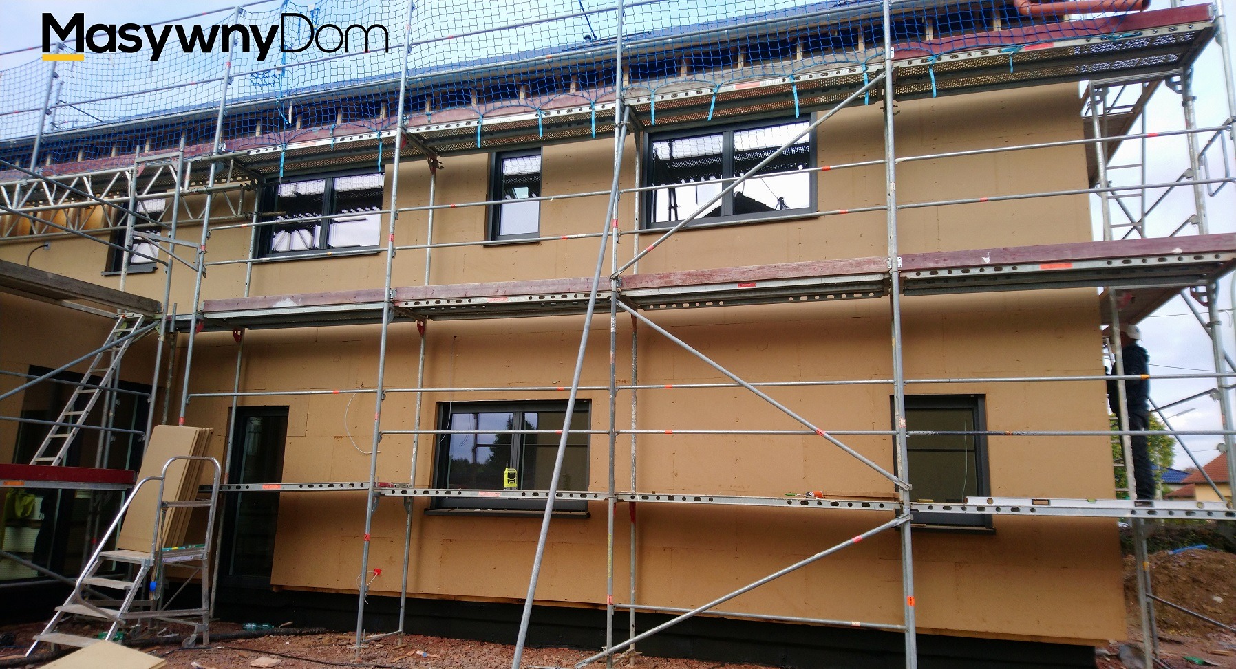 Budowa domu w trakcie ocieplania, widoczne rusztowania, czarne okna i siatka zabezpieczająca na górze