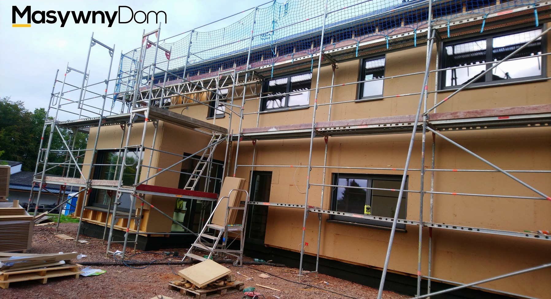 Budowa domu w technologii szkieletowej z widocznym rusztowaniem, siatką zabezpieczającą na dachu i zamontowanymi oknami, na placu budowy leżą materiały.