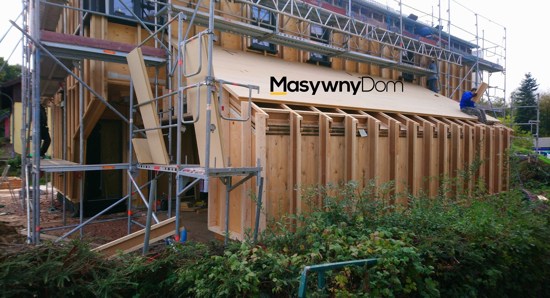 Konstrukcja domu w trakcie budowy, widoczne drewniane elementy szkieletu, rusztowania i pracownicy na dachu, z logo firmy na elewacji.