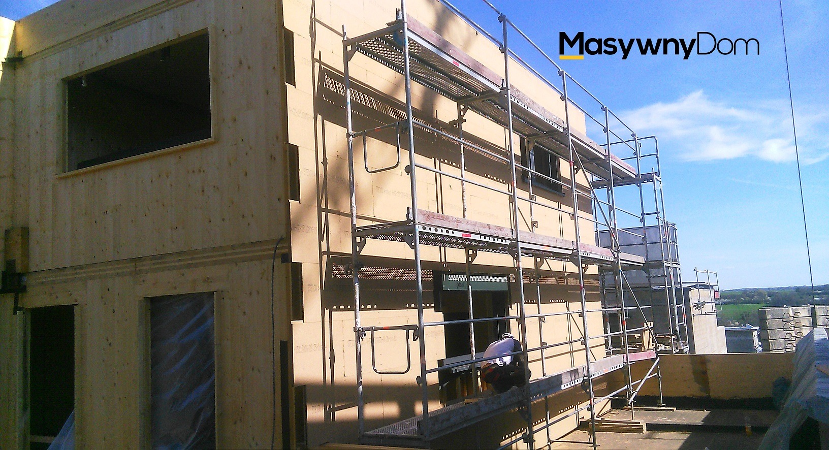 Wznoszenie dwukondygnacyjnego domu z drewnianych paneli, widoczne rusztowania i pracownik w kasku na tle błękitnego nieba.