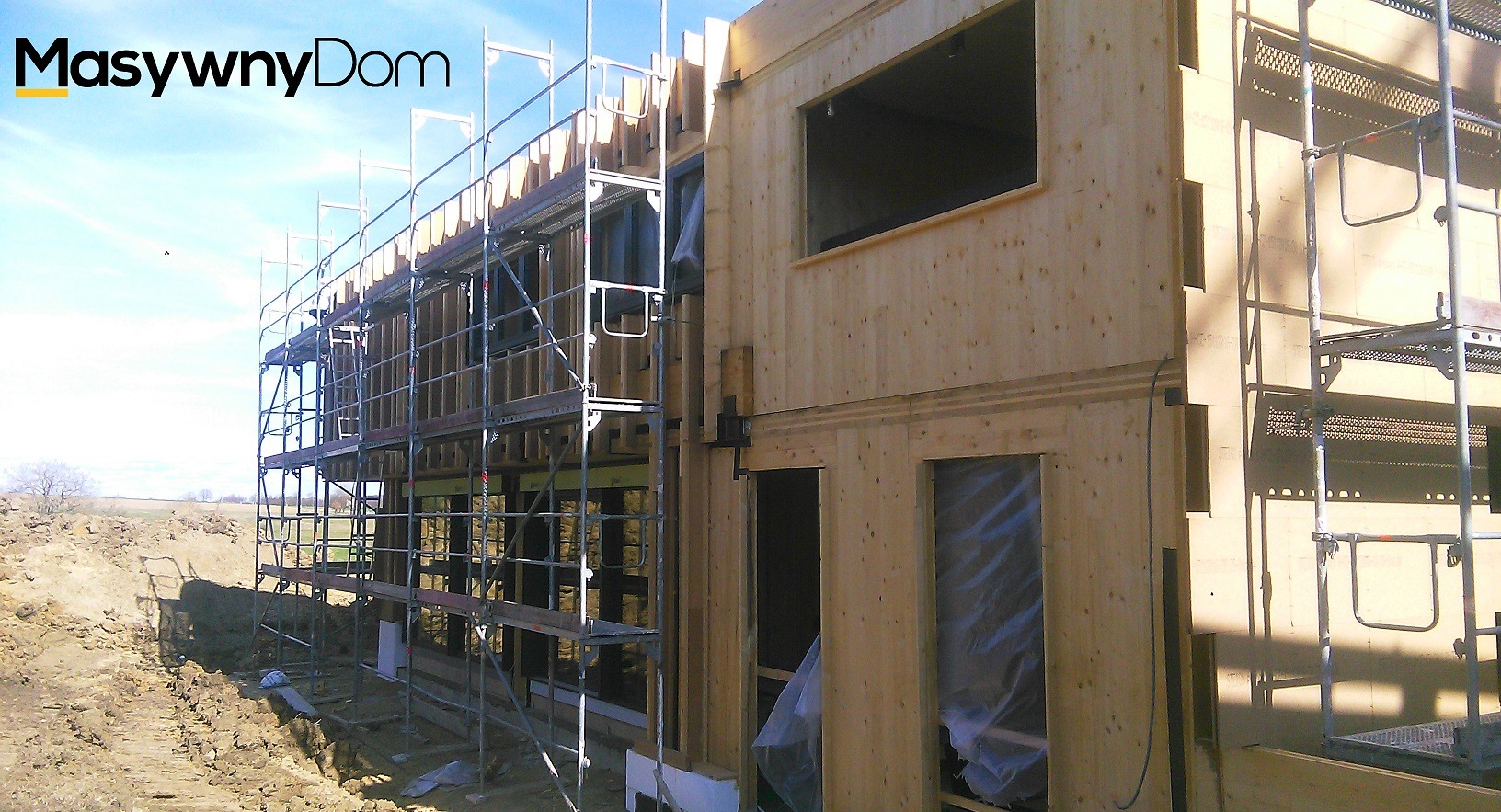 Wznoszenie dwukondygnacyjnego domu szkieletowego z drewnianych paneli, z widocznymi rusztowaniami i wykopem pod fundamenty.
