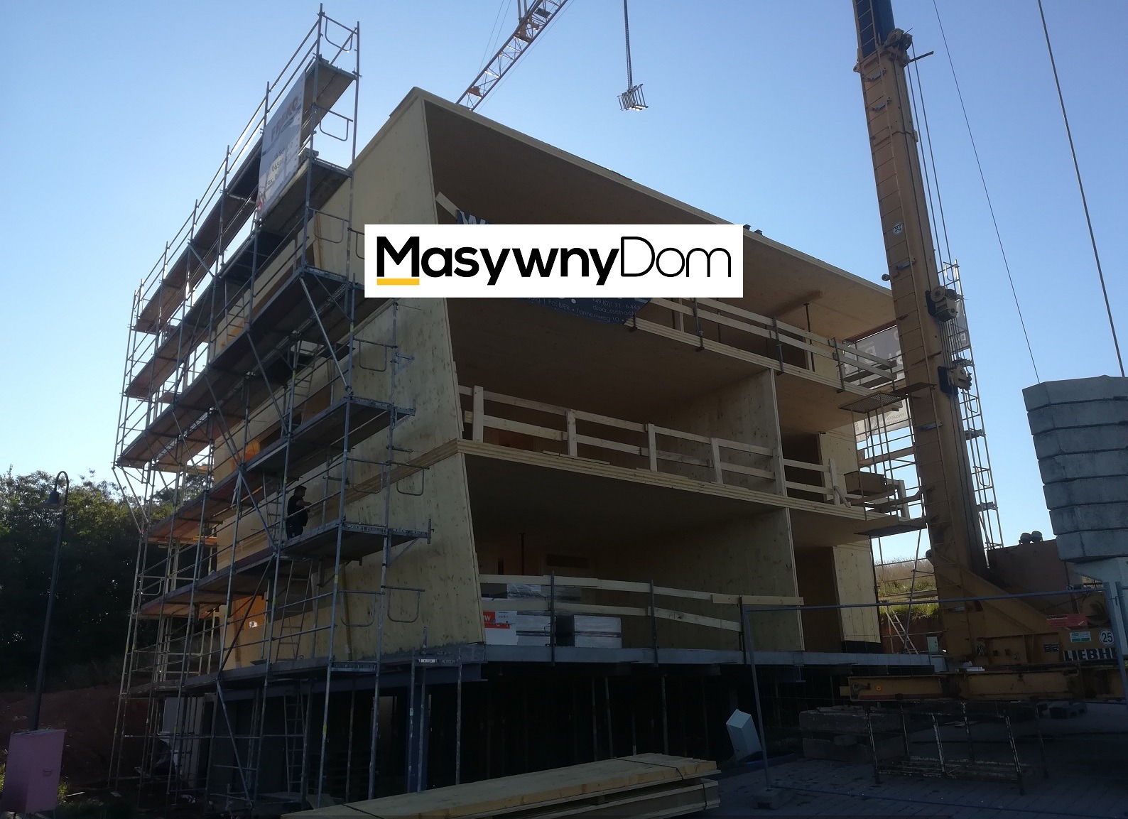 Wznoszenie trzypiętrowego domu w konstrukcji szkieletowej z wykorzystaniem rusztowań i dźwigu, widoczne płyty warstwowe i drewniane elementy konstrukcyjne.