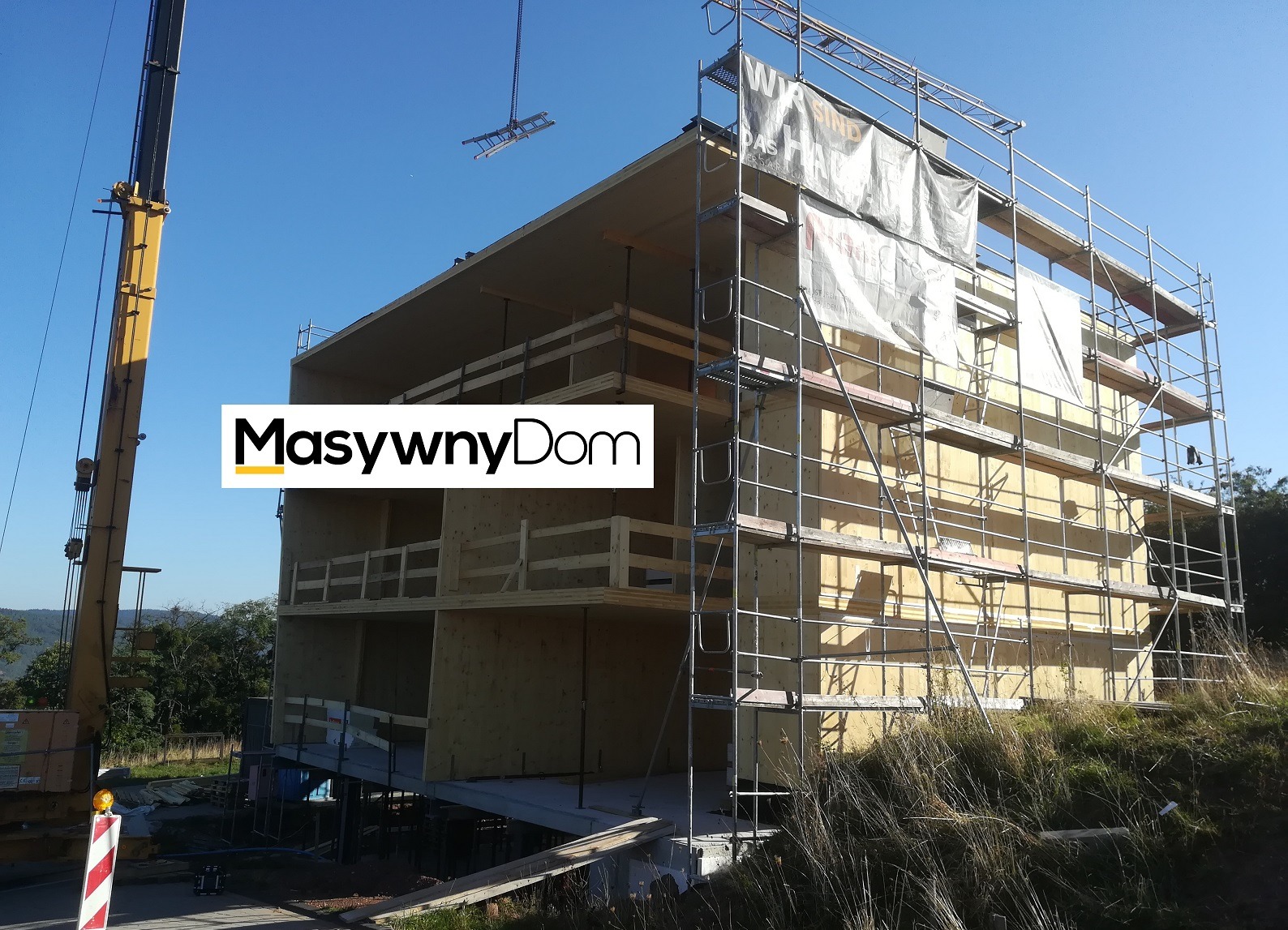 Wznoszenie dwukondygnacyjnego domu szkieletowego z drewnianą konstrukcją, widoczne rusztowania, dźwig z podwieszonymi schodami i banery reklamowe na fasadzie w słoneczny dzień.
