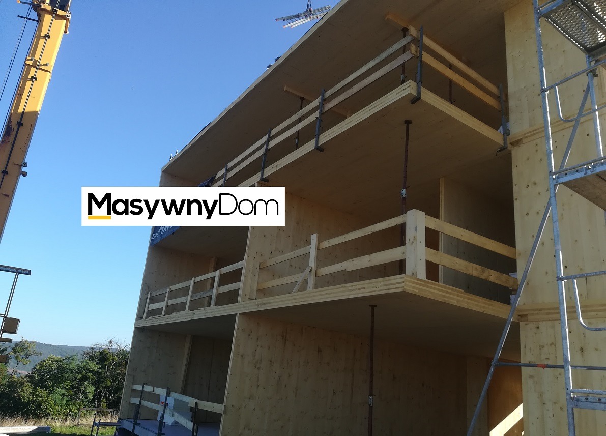 Wielopiętrowy dom szkieletowy w trakcie budowy, widoczne drewniane elementy konstrukcyjne, zabezpieczenia balkonowe, żuraw budowlany i rusztowanie, błękitne niebo w tle.