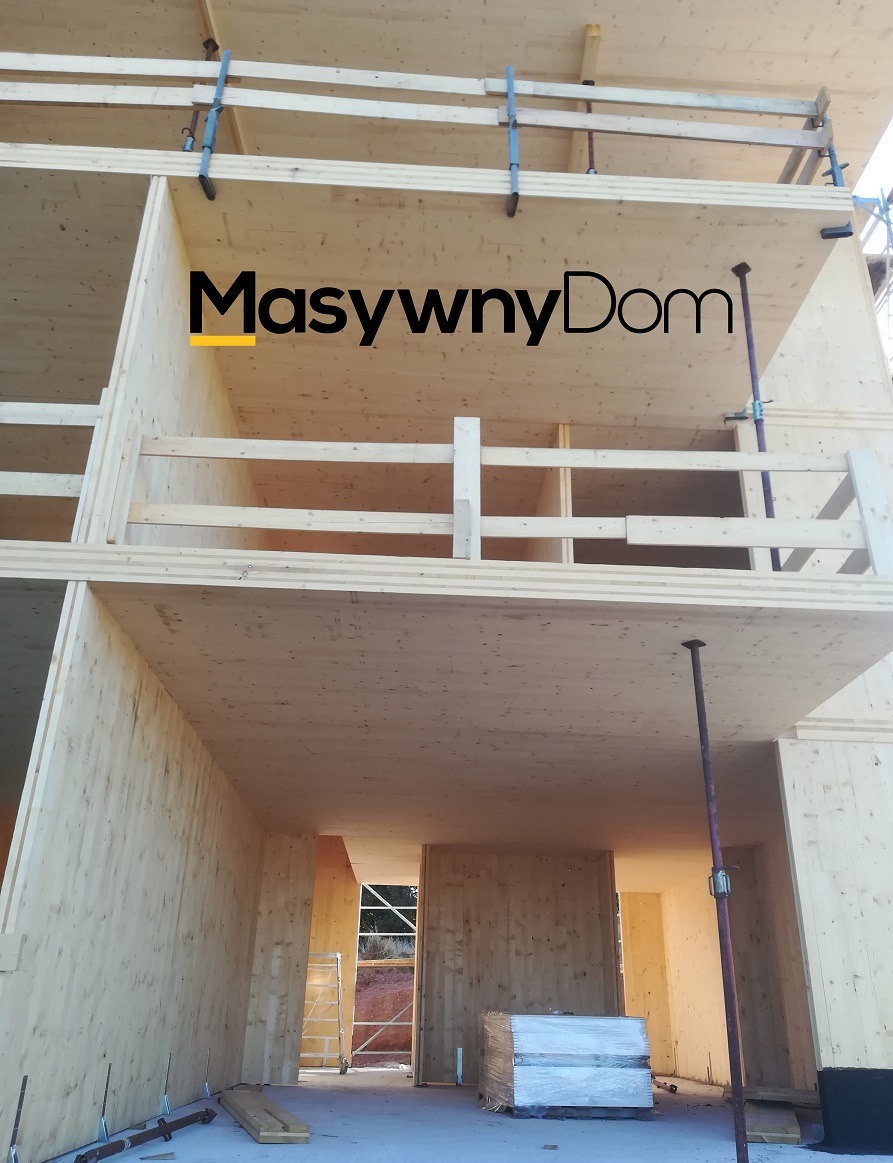 Wielopiętrowa konstrukcja w trakcie budowy, drewniane ściany i stropy, widoczne rusztowania i zabezpieczenia, logo firmy na elewacji, podłoże wyłożone folią.