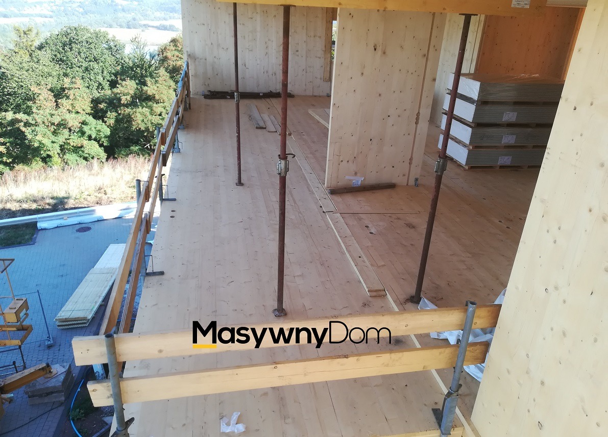 Wnętrze w trakcie budowy, widoczne drewniane ściany i podłoga, podpory stalowe, paczki płyt na paletach, zabezpieczenia na krawędzi dachu i otoczenie z drzewami w tle.