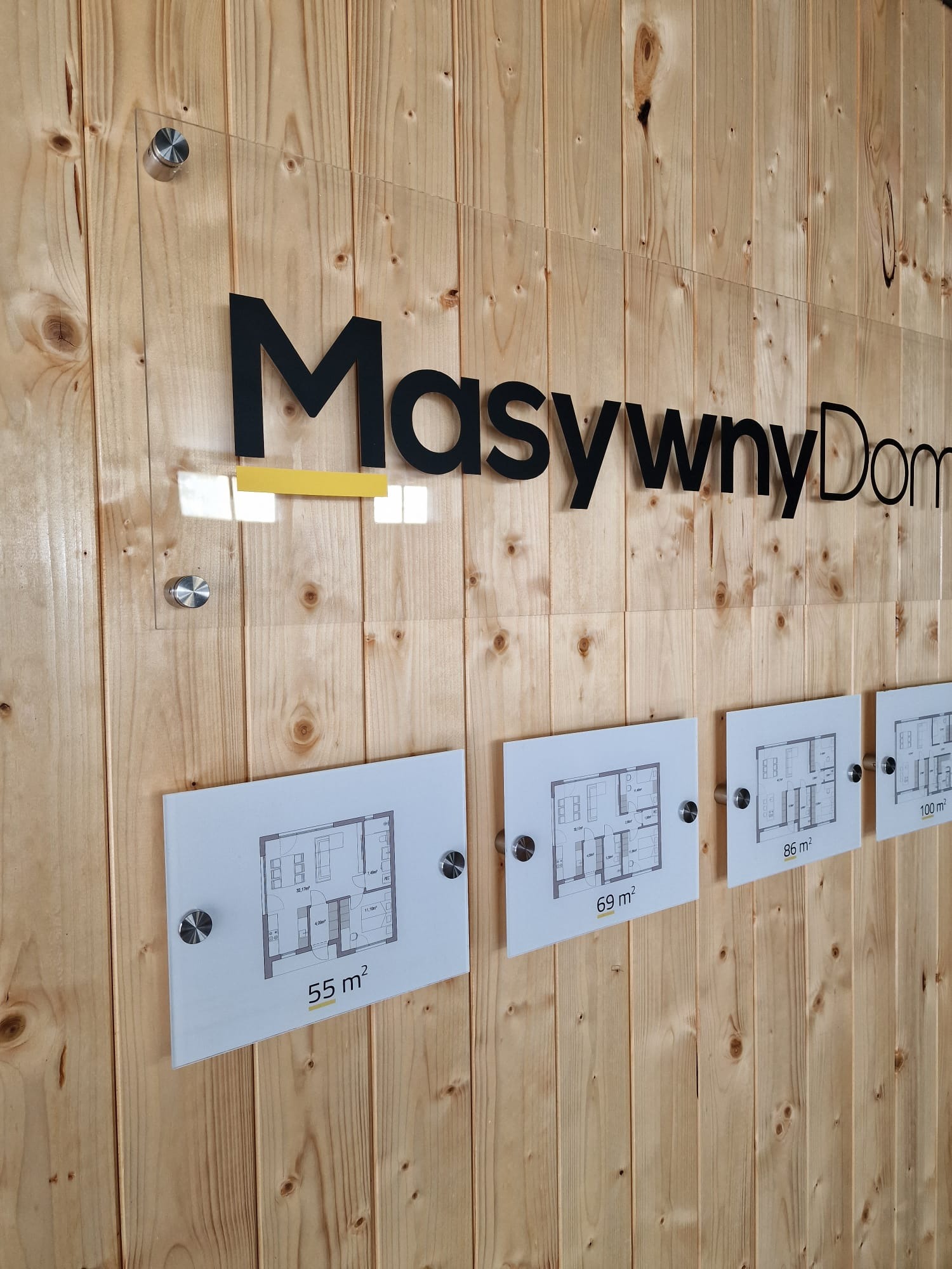 Ekspozycja firmy budowlanej Masywny Dom na drewnianej ścianie z prezentacją planów domów o powierzchniach 55, 69, 86 i 100 metrów kwadratowych.