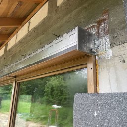 Majgo Łukasz Rudzki - Montaż zewnętrznej rolety z aluminiową obudową marki Aluprof nad oknem z drewnianą ramą, widoczne elementy konstrukcyjne budynku i warstwa izolacyjna